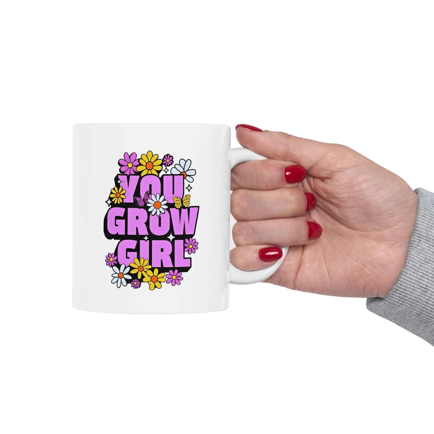 Hand holding a white ceramic mug featuring the text ‘YOU GROW GIRL’ in bold purple letters surrounded by colorful flowers.