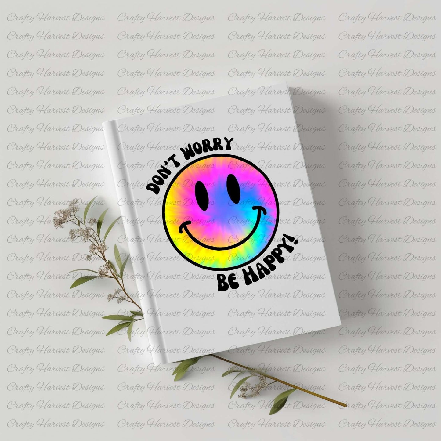 White hardcover notebook featuring a vibrant tie-dye smiley face design with the phrase ‘Don’t Worry, Be Happy!’ in black text. The notebook is placed on a light-colored surface with dried plant stems beside it, creating a soft and aesthetic composition.