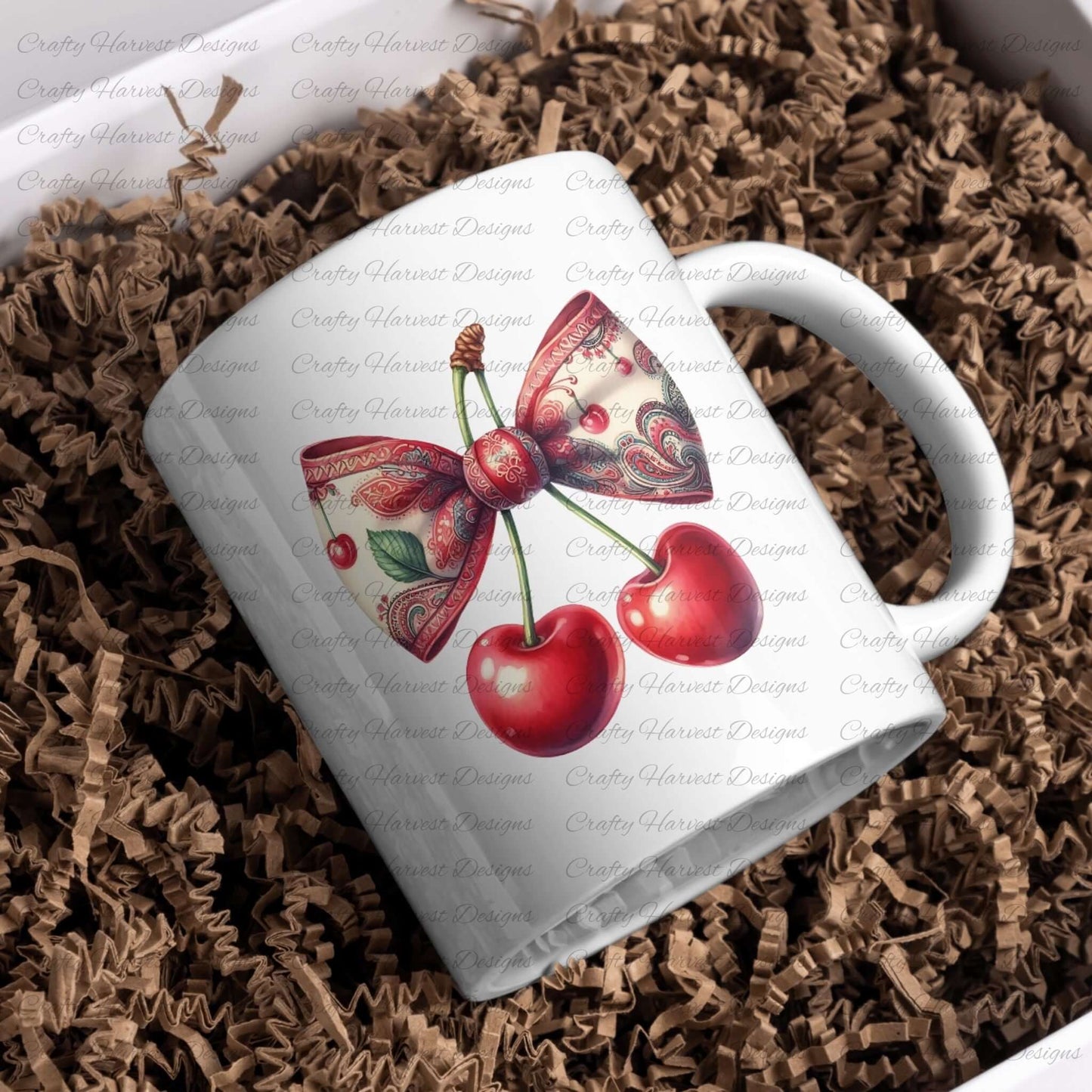 A white ceramic mug featuring a cherry design with a paisley-patterned bow in red and green tones. The mug is nestled in brown crinkle paper, giving it a cozy presentation.