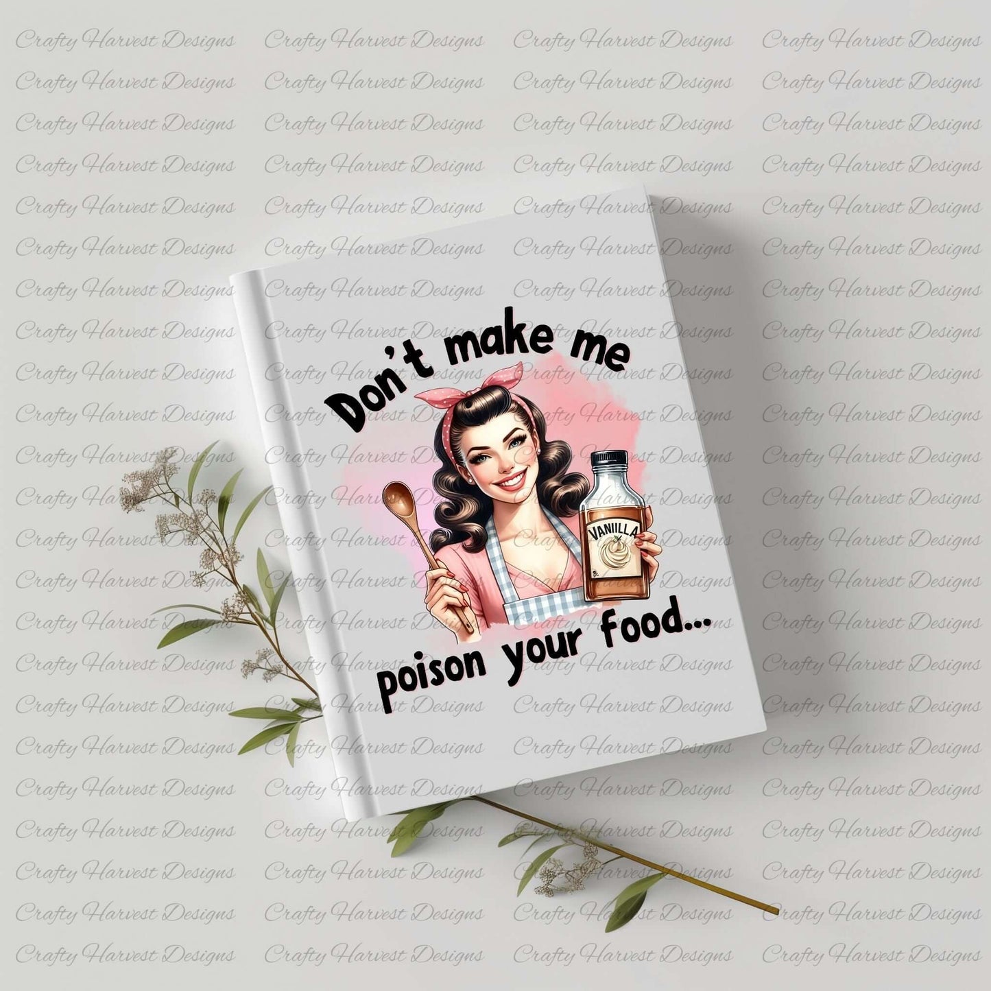 A white notebook with a vintage-style design on the cover featuring a smiling retro housewife holding a wooden spoon and a bottle labeled “Vanilla.” The text above and below the illustration reads, “Don’t make me poison your food…” The notebook is placed on a light surface with green foliage nearby.