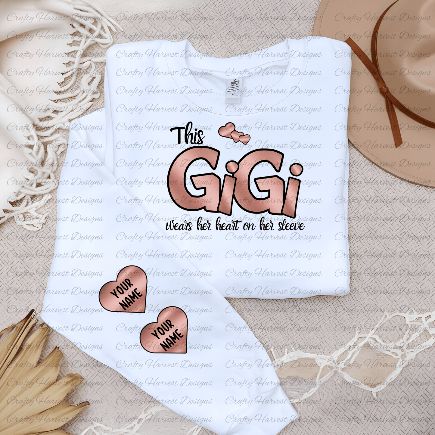 Mockup of a white t-shirt featuring the “This GiGi Wears Her Heart on Her Sleeve” design in a brown color variation. The shirt also displays two customizable heart accents on the sleeve with the placeholder text “Your Name.” The image includes neutral decor elements like a hat and woven fabric for a cozy, styled look.
