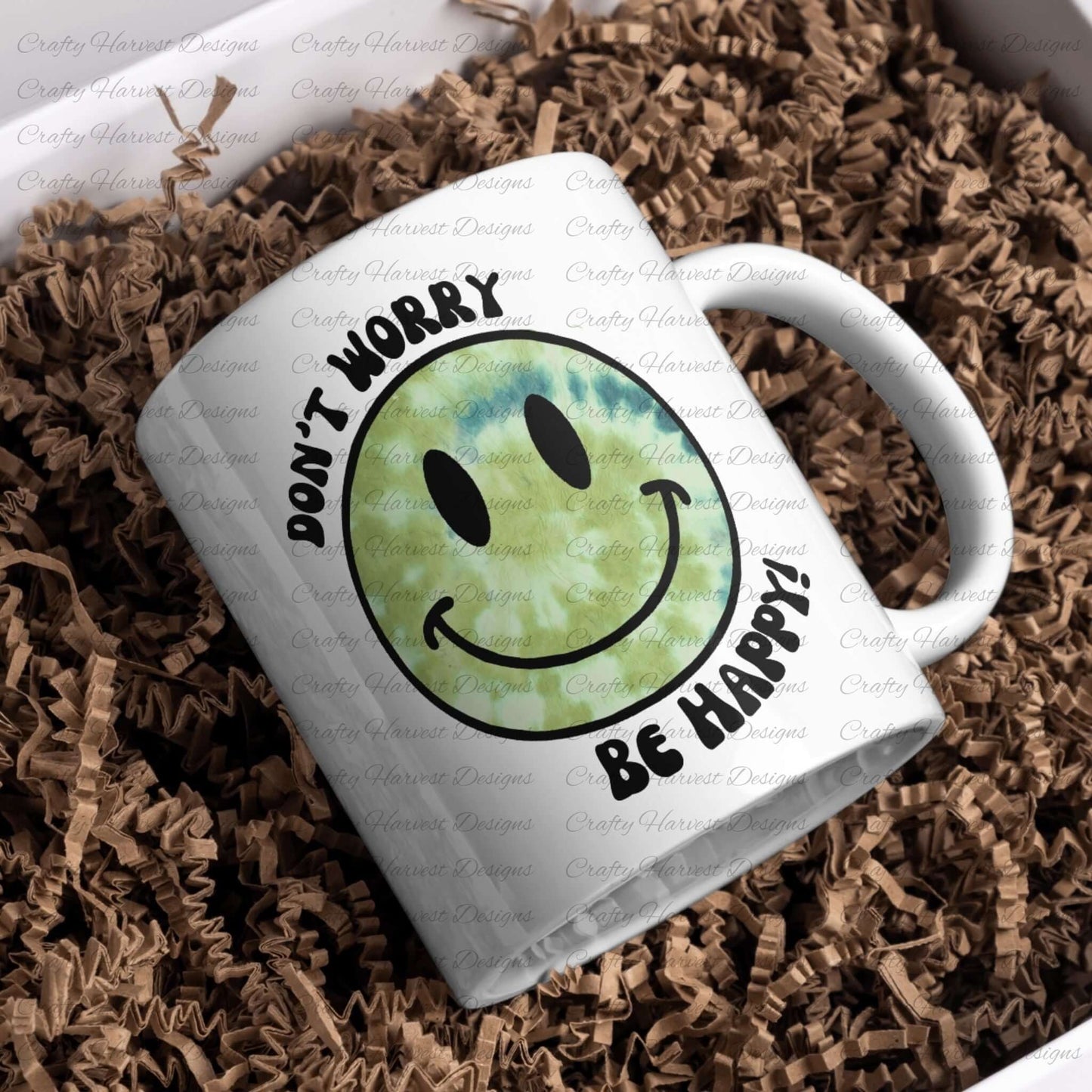 White ceramic mug featuring a green tie-dye smiley face design with the phrase ‘Don’t Worry, Be Happy!’ in black text. The mug is placed on a bed of crinkled brown paper filler.