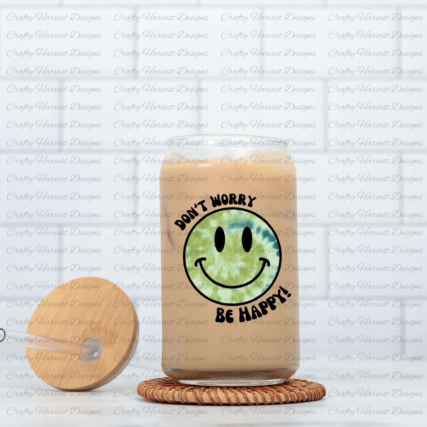 Glass can tumbler filled with iced coffee, featuring a green tie-dye smiley face design with the phrase ‘Don’t Worry, Be Happy!’ in black text. The tumbler is placed on a woven coaster, with a wooden lid and glass straw resting beside it.
