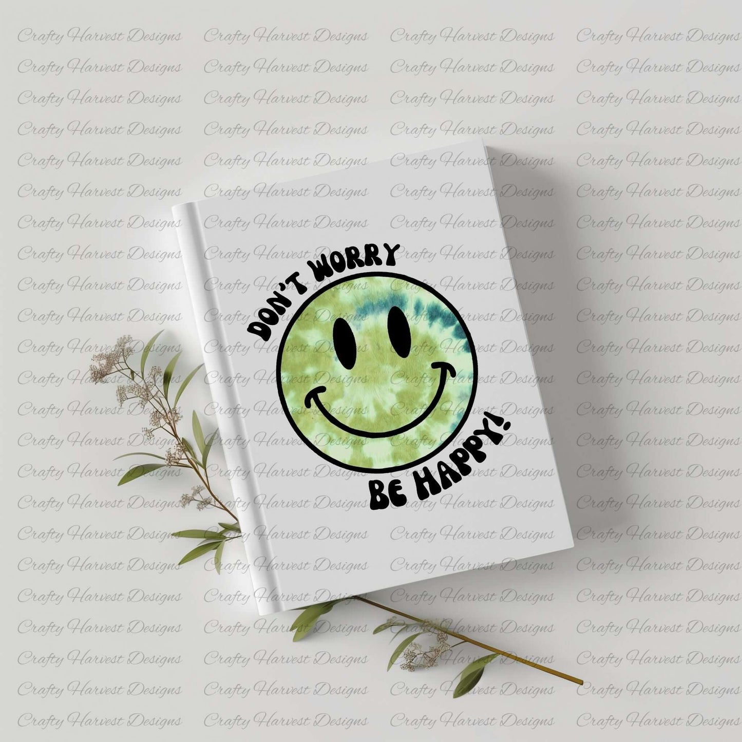White hardcover notebook featuring a green tie-dye smiley face design with the phrase ‘Don’t Worry, Be Happy!’ in black text. The notebook is placed on a light-colored surface with dried plant stems beside it.