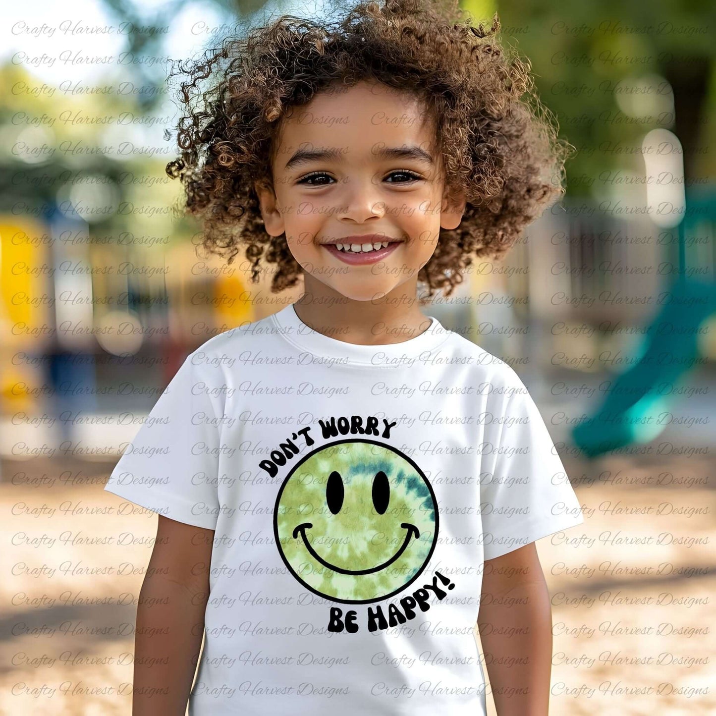 Young child wearing a white t-shirt featuring a green tie-dye smiley face with “Don’t Worry, Be Happy!” text. The child is smiling outdoors.