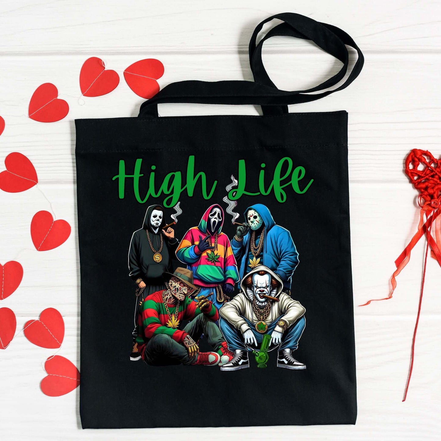 Black tote bag featuring the ‘High Life’ design with five masked characters in streetwear holding cannabis-themed accessories. The text ‘High Life’ is displayed in a bold green script font above the graphic. The bag is placed on a white wooden surface with red paper hearts and ribbon decorations around it.