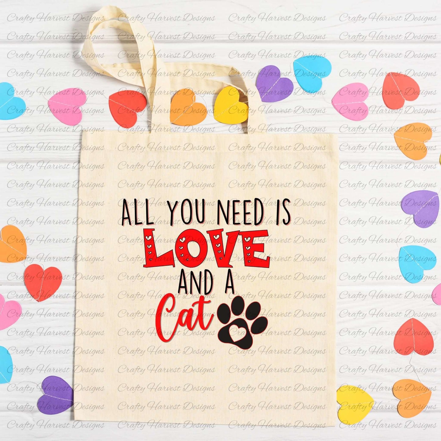 Canvas tote bag with the All You Need is Love and a Cat design in red and black with a paw print graphic, surrounded by colorful paper hearts on a white wooden background.