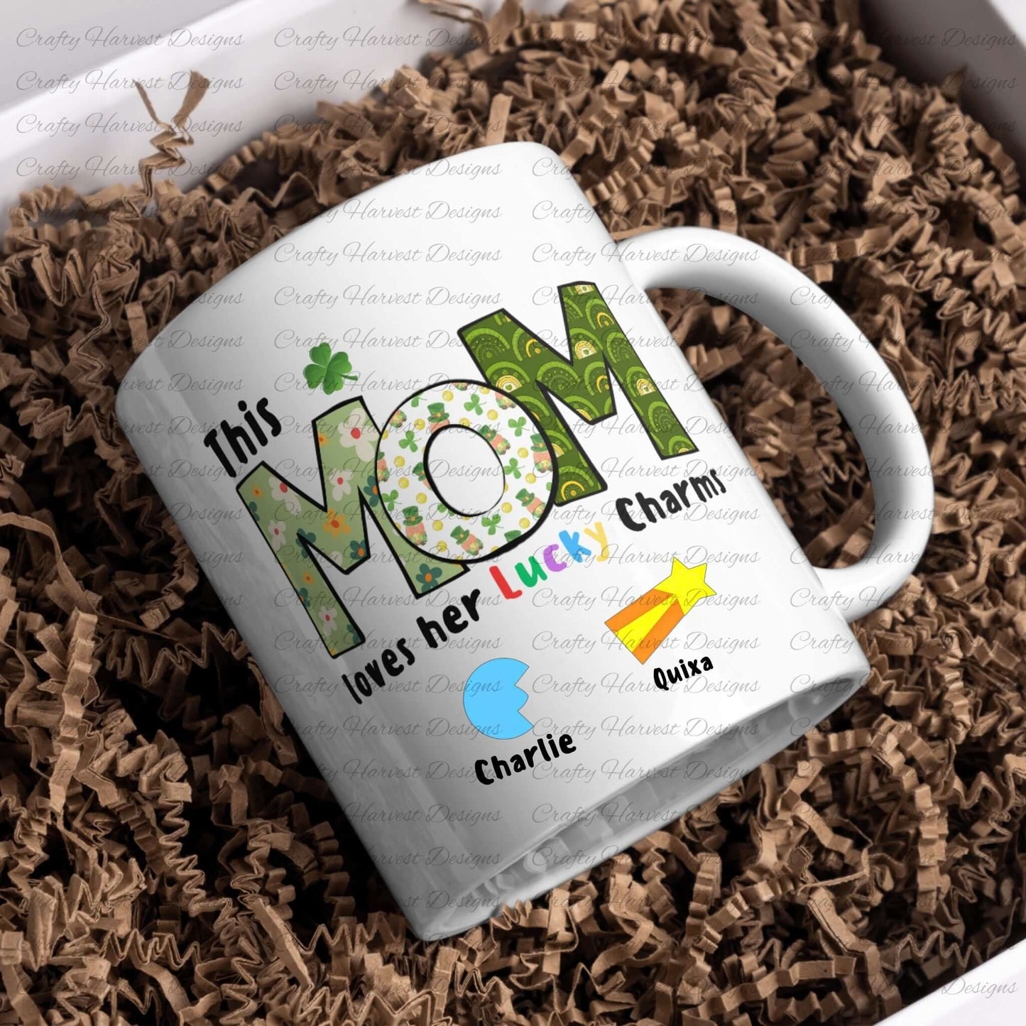 A white ceramic mug featuring the design “This MOM loves her Lucky Charms” with patterned green letters and a shamrock above the “O.” Below the text are two colorful lucky charm icons (a blue crescent and a yellow shooting star) with the names “Charlie” and “Quixa” beneath each charm. The mug is placed on brown crinkle paper.
