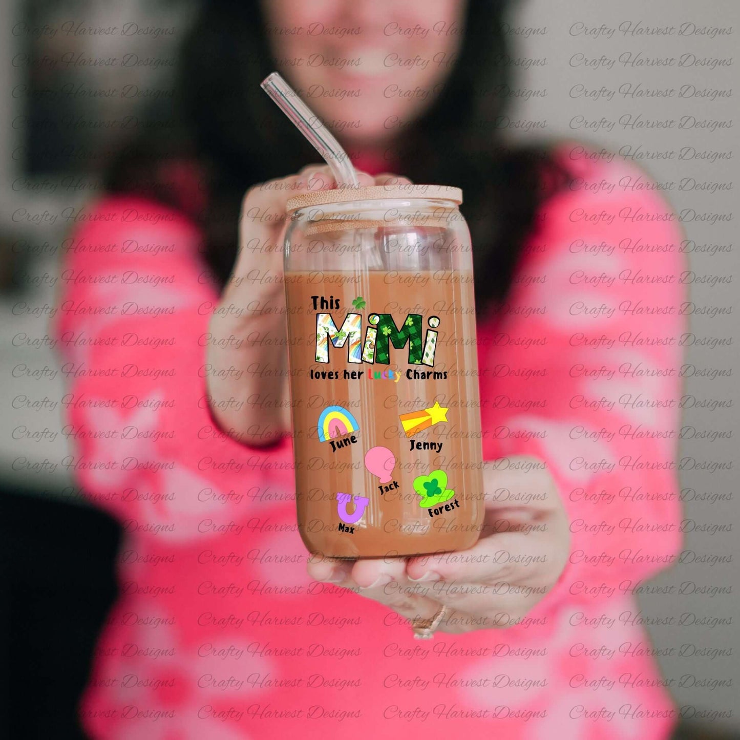 A person wearing a bright pink shirt holds a glass can with a bamboo lid and glass straw. The design on the glass reads “This MIMI loves her Lucky Charms” with green-patterned letters and a shamrock above the “I.” Below the text are six colorful lucky charm icons (rainbow, pink balloon heart, horseshoe, shooting star, and clover) with the names “June,” “Max,” “Jenny,” and “Forest” next to them. The glass is filled with an iced coffee or similar drink.