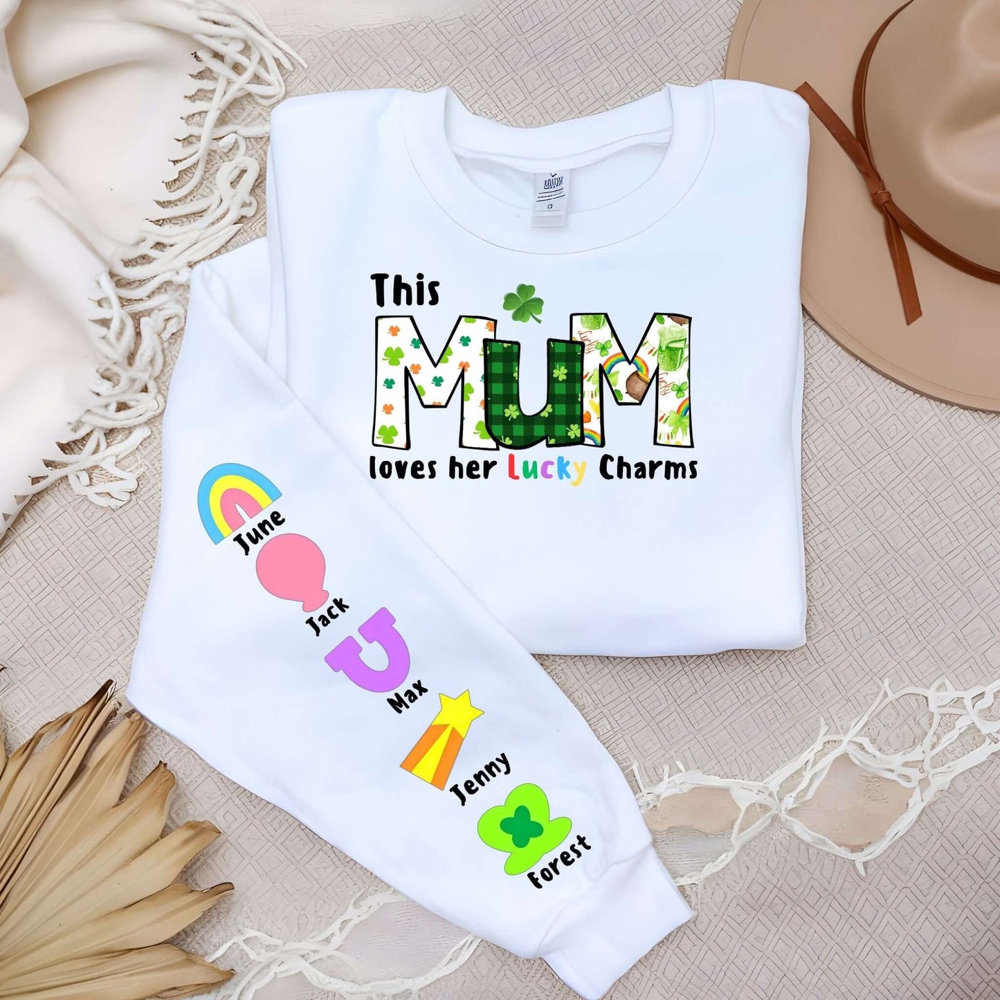 A white long-sleeve shirt featuring the design “This MUM loves her Lucky Charms” with patterned green and floral letters, a shamrock above the “U,” and colorful text. The left sleeve displays six colorful lucky charms (rainbow, balloon heart, horseshoe, shooting star, and clover) with names “June,” “Jack,” “Max,” “Jenny,” and “Forest” beside each charm. The shirt is laid on a neutral fabric with decorative palm leaves and a tan hat nearby.