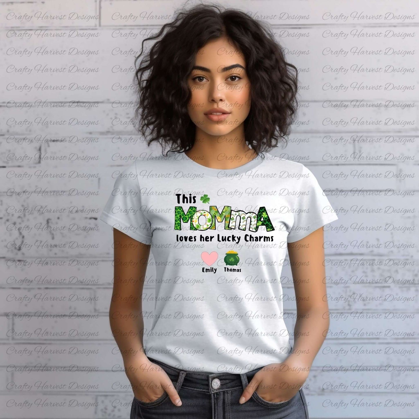 A woman wears a white t-shirt featuring the design “This MOMMA loves her Lucky Charms” with green-patterned letters and a shamrock above the “M.” Below the text are two colorful lucky charm icons (a pink heart and a green clover) with the names “Emily” and “Thomas” underneath. The background is a light gray brick wall.