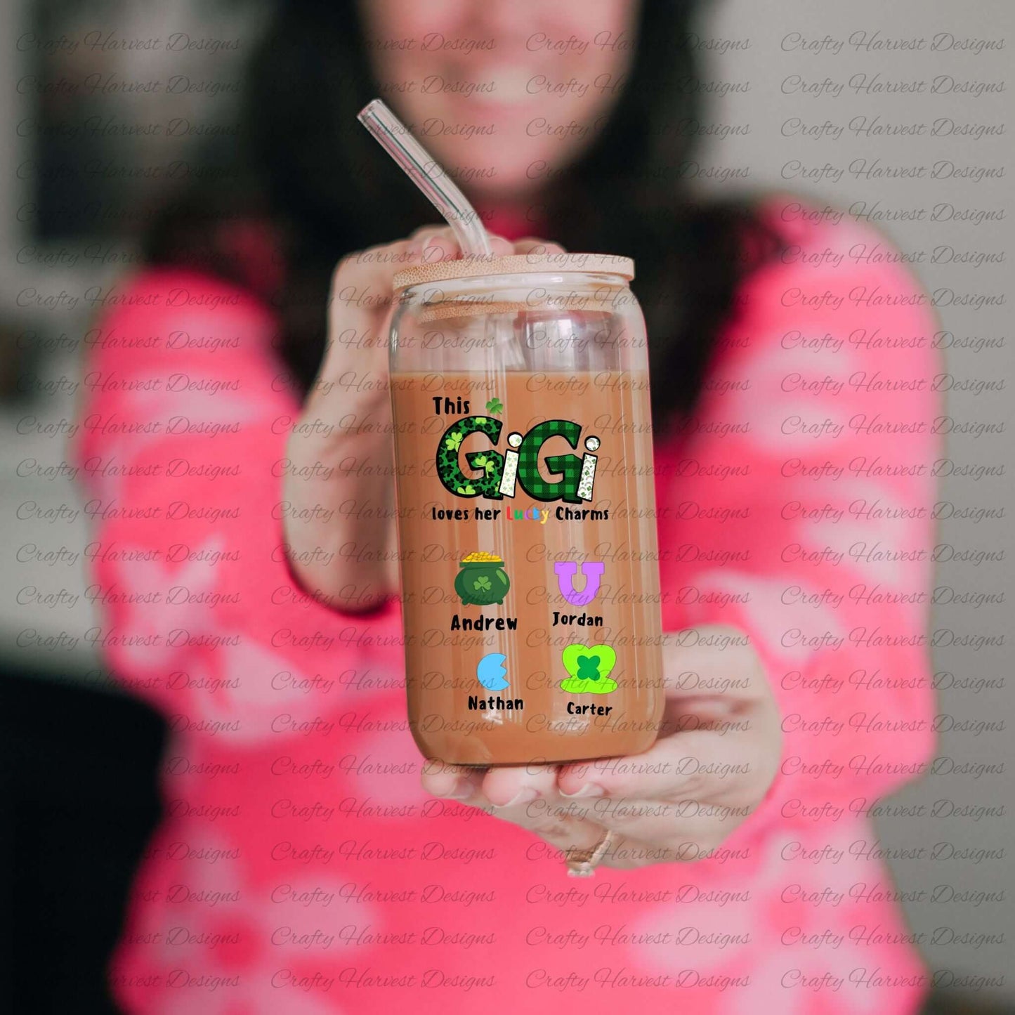 Person holding a glass can with a bamboo lid and straw, featuring a “This GIGI loves her Lucky Charms” design with green-patterned letters and two colorful charm icons with the names “Emily” and “Thomas” below them.