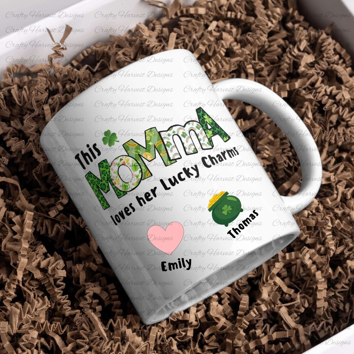 White ceramic mug with a “This MOMMA loves her Lucky Charms” design, featuring green-patterned letters and two colorful charm icons with the names “Emily” and “Thomas” below them.