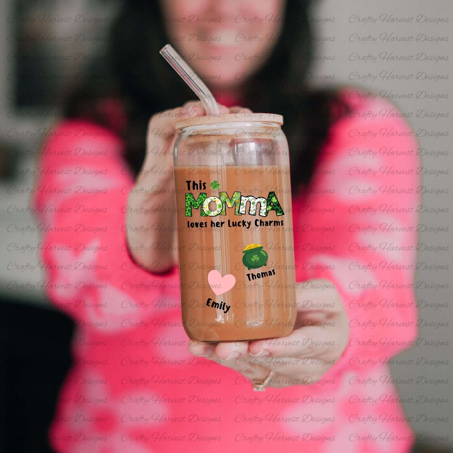 Person holding a glass can with a bamboo lid and straw, featuring a “This MOMMA loves her Lucky Charms” design with green-patterned letters and two colorful charm icons with the names “Emily” and “Thomas” below them.