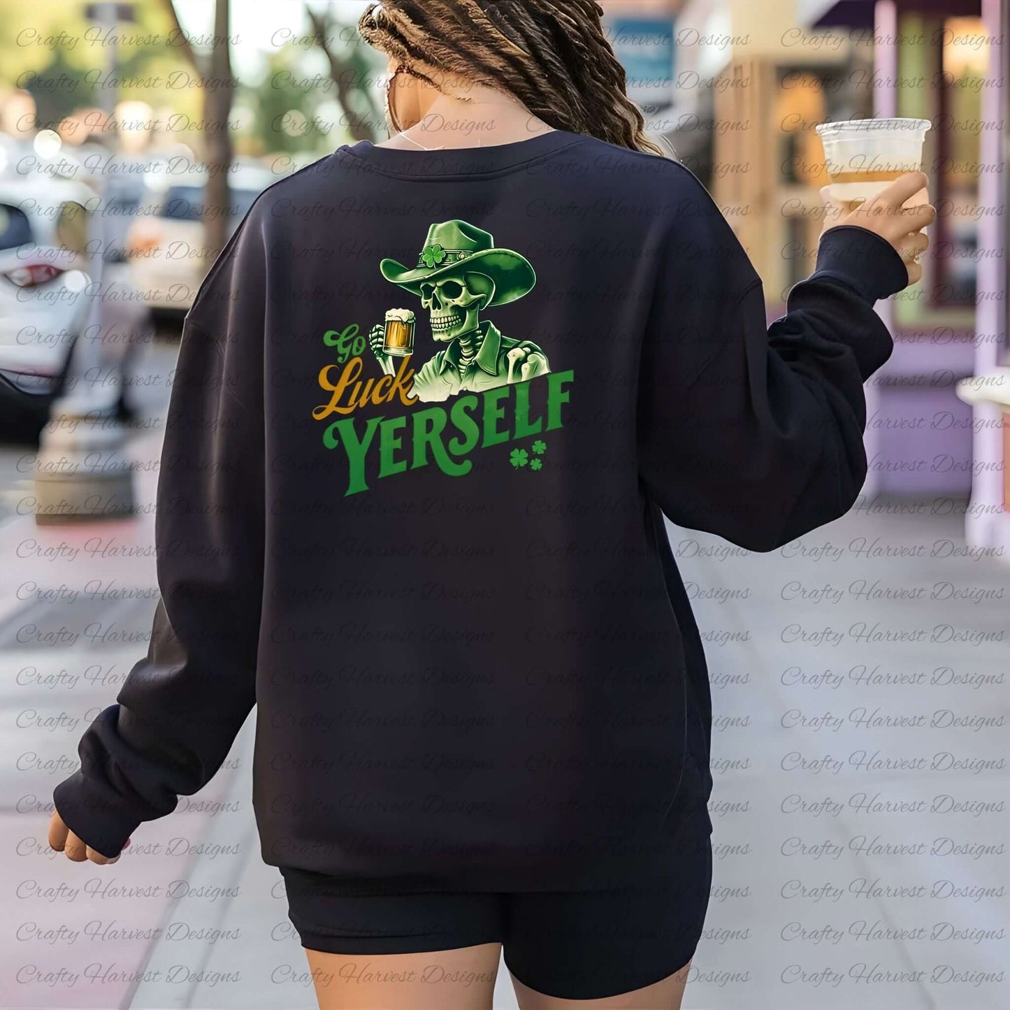 A person is seen from the back, holding a clear plastic cup with a drink in their right hand, while wearing a black sweatshirt. The sweatshirt features a graphic of a skeleton in a green cowboy hat with a shamrock, holding a beer mug. The text on the design reads “Go Luck Yourself” in green and yellow fonts with two small shamrocks beneath the text. The person is walking outdoors on a colorful city street