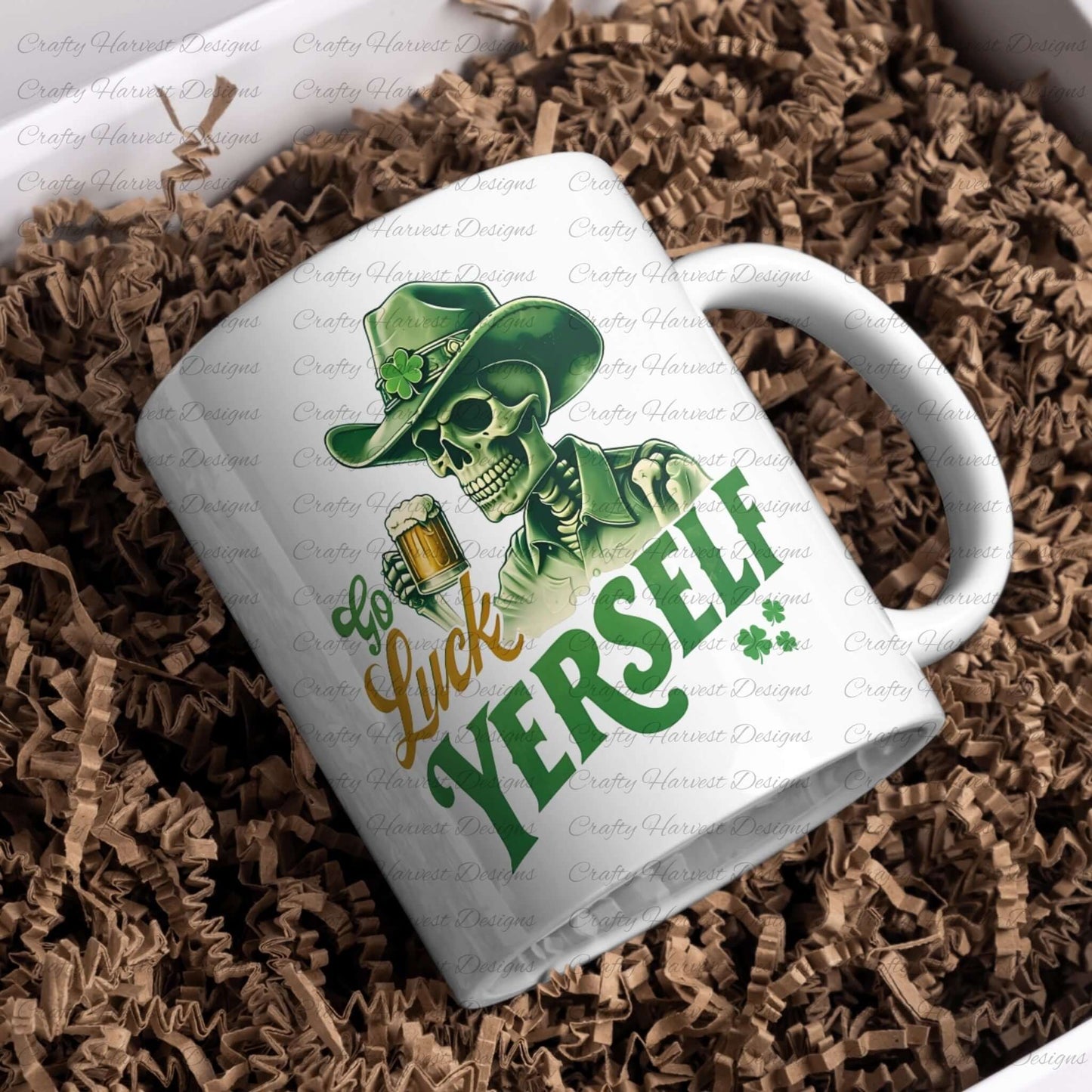 A white ceramic mug rests on a bed of crinkled brown paper filler. The mug features a graphic design of a skeleton wearing a green cowboy hat adorned with a shamrock, holding a beer mug. The text on the mug reads “Go Luck Yourself” in green and yellow fonts with two small shamrocks below the text.