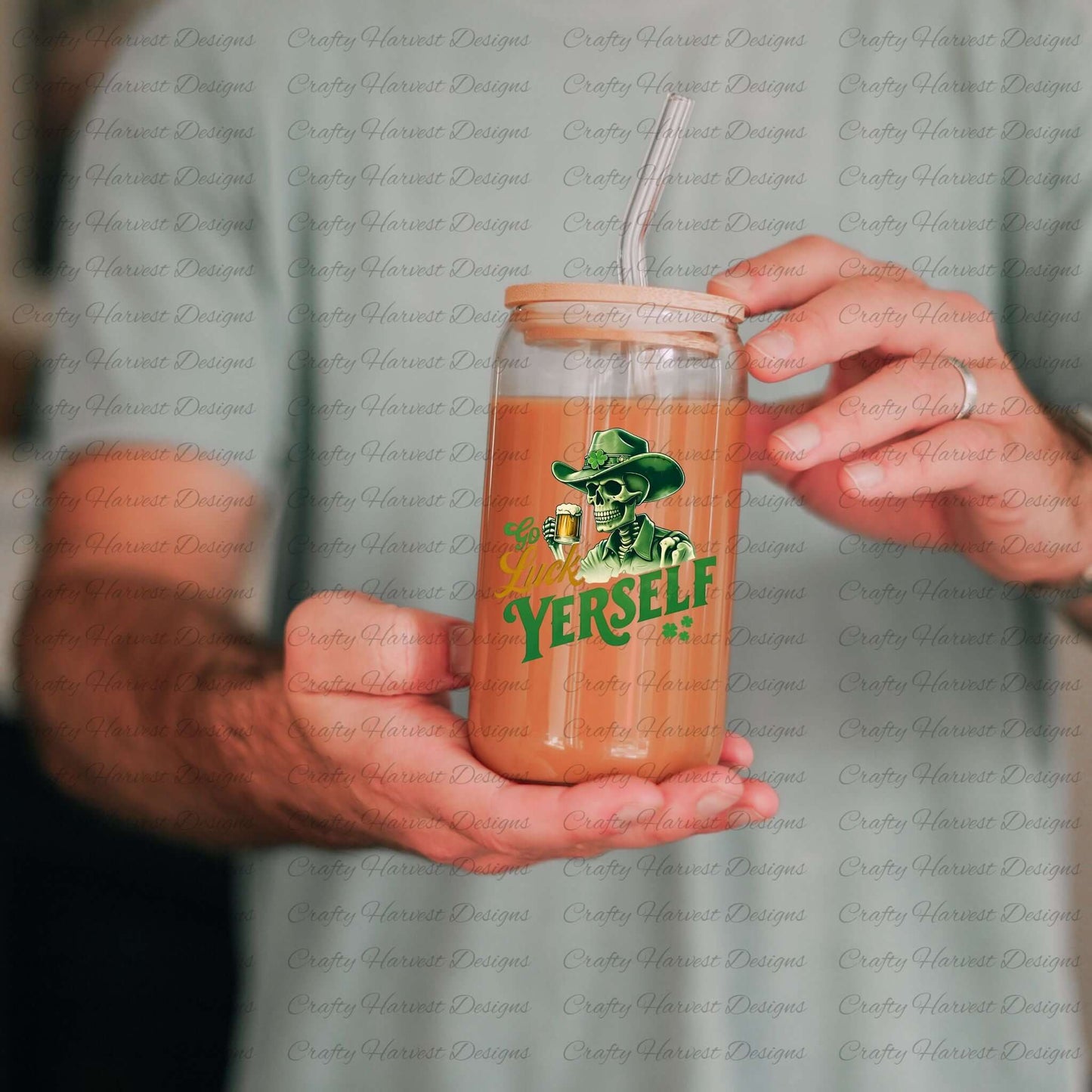 A person wearing a light-colored shirt holds a glass can with a bamboo lid and metal straw. The glass can contains an iced coffee drink and features a green and yellow graphic design of a skeleton wearing a cowboy hat adorned with a shamrock, holding a beer mug. The text reads “Go Luck Yourself” with two small shamrocks below the text.
