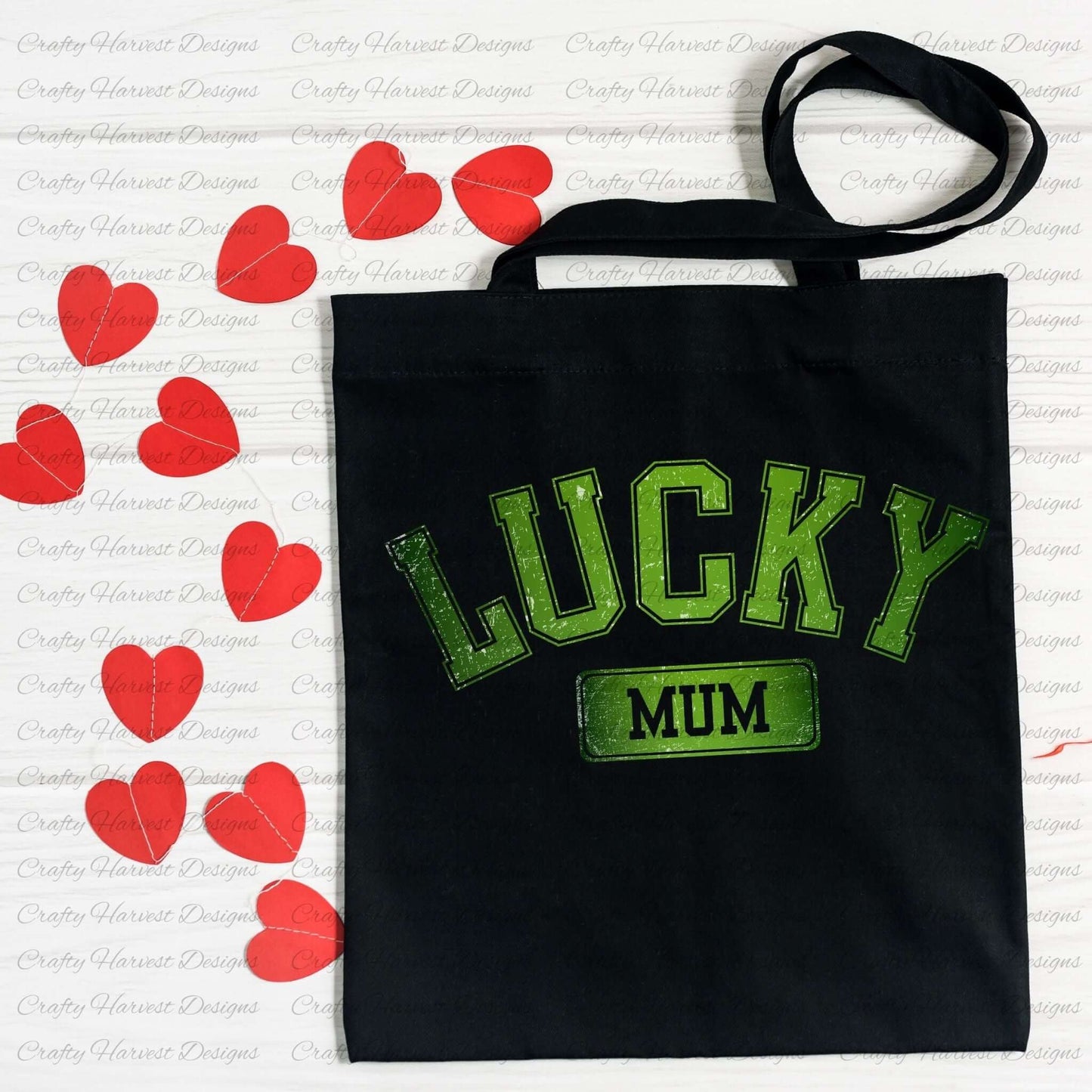 Black tote bag featuring the design ‘Lucky Mum’ in bold green text with a St. Patrick’s Day theme. The bag is displayed on a surface scattered with red heart decorations.