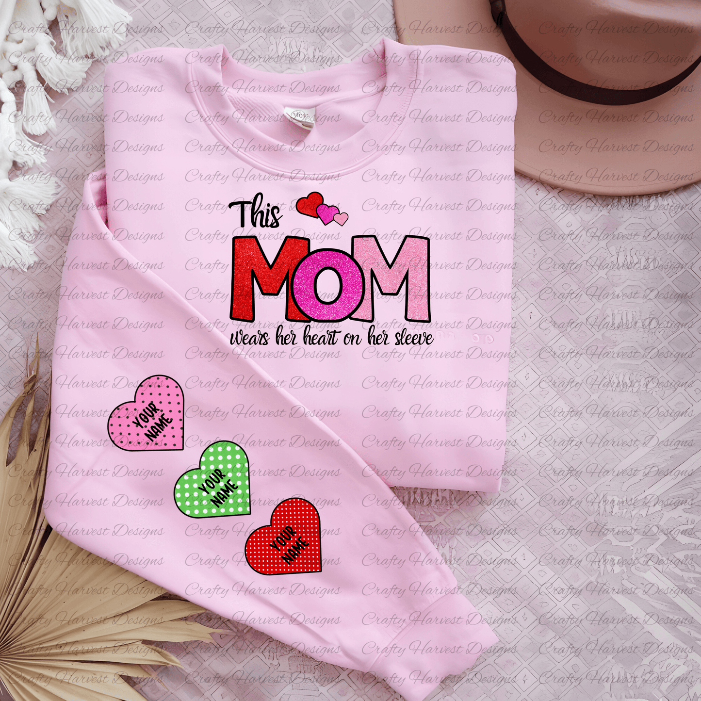 Mockup of a pink t-shirt featuring the “This Mom Wears Her Heart on Her Sleeve” design in a glittery red and pink color variation. The sleeve displays three customizable heart accents in pink polka dot, green polka dot, and red polka dot patterns with the placeholder text “Your Name.” The shirt is styled with neutral decor elements for a cozy look.