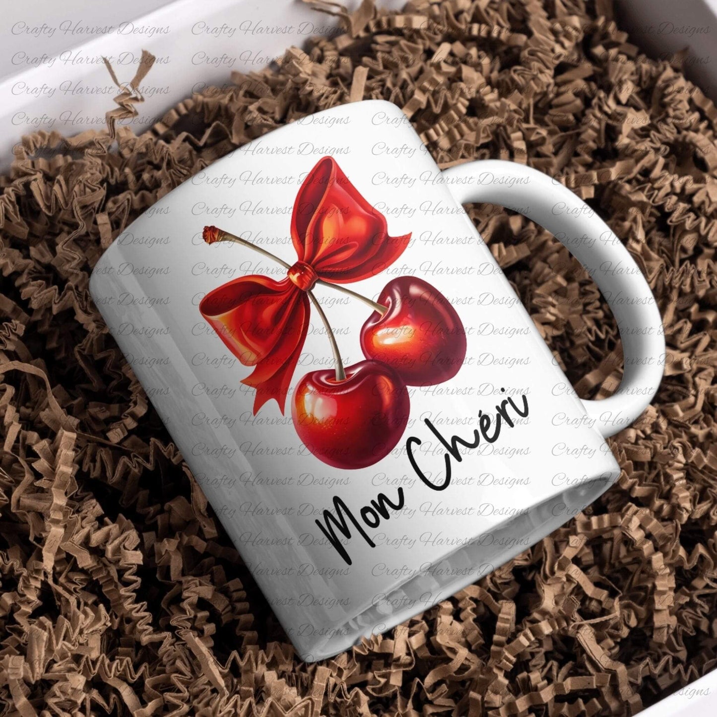 White ceramic mug featuring a digital design of two vibrant red cherries tied with a bold red bow, accompanied by the elegant black text ‘Mon Chéri.’ The mug is nestled in brown shredded paper, showcasing its appeal as a stylish and customizable drinkware item or gift.