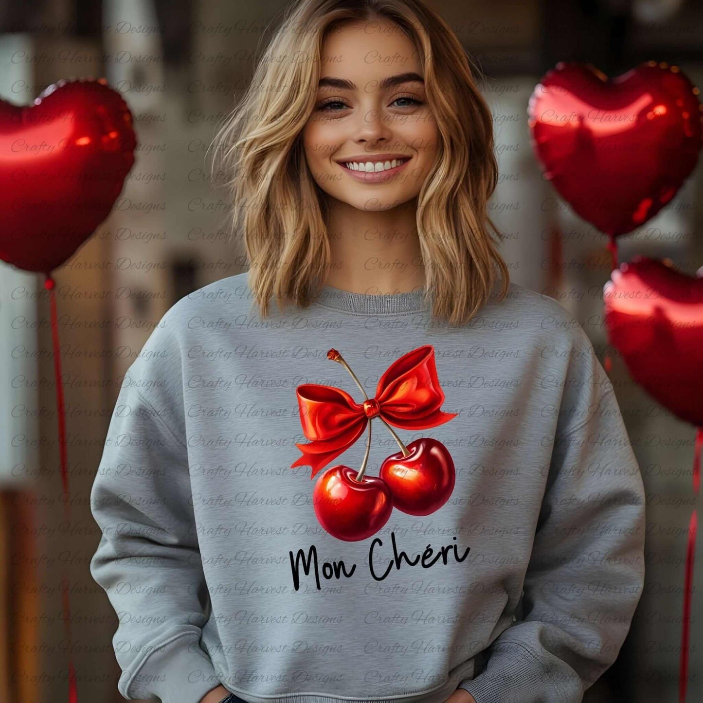 Smiling woman wearing a gray sweatshirt featuring a digital design of two red cherries tied with a bold red bow, accompanied by the elegant black text ‘Mon Chéri.’ Red heart-shaped balloons in the background enhance the romantic and playful theme of the design.