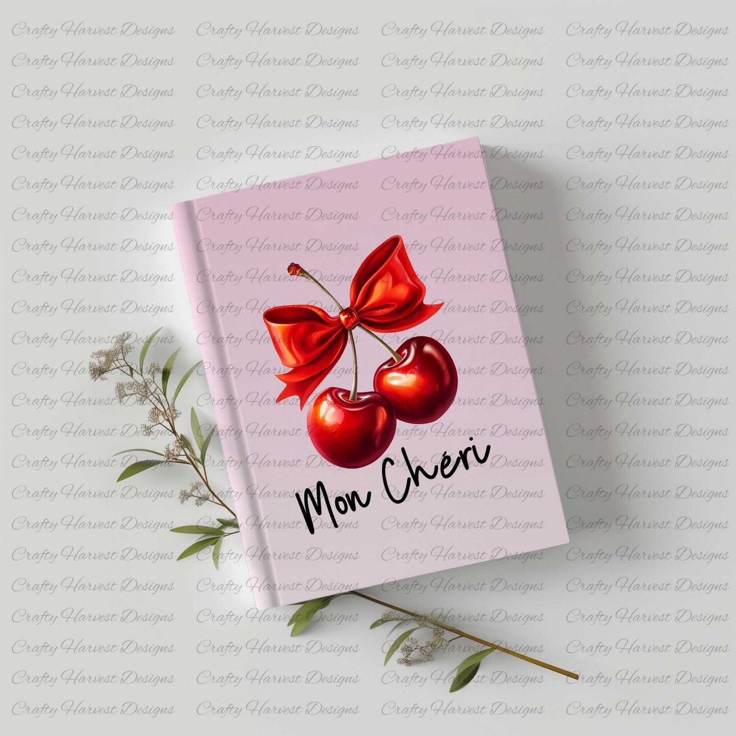 Pink hardcover notebook featuring a digital design of two red cherries tied with a bold red bow, accompanied by the elegant black text ‘Mon Chéri.’ The notebook is displayed on a light surface with sprigs of greenery, highlighting its use as a stylish and functional stationery item.