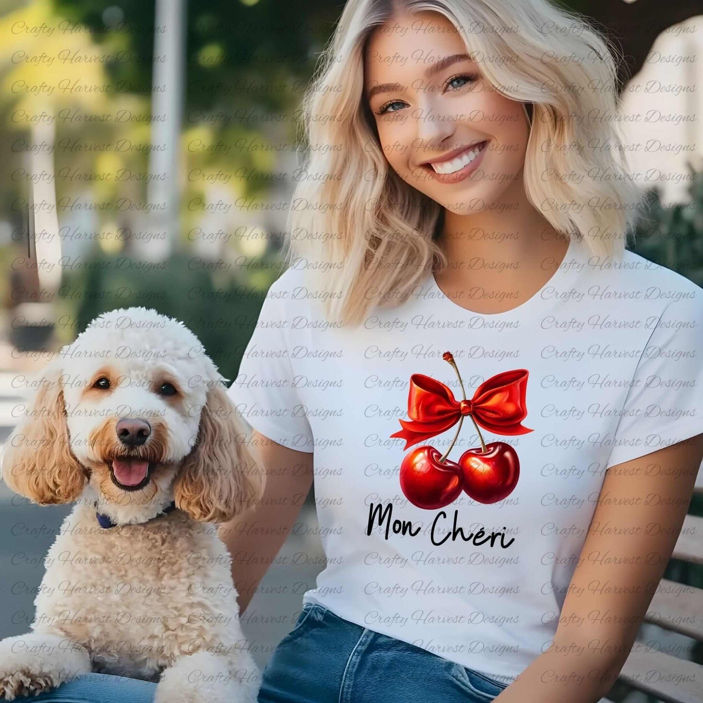 Smiling woman wearing a white t-shirt featuring a digital design of two red cherries tied with a bold red bow, accompanied by the elegant black text ‘Mon Chéri.’ She is seated outdoors with a fluffy white dog, showcasing the design as a stylish and playful apparel option.