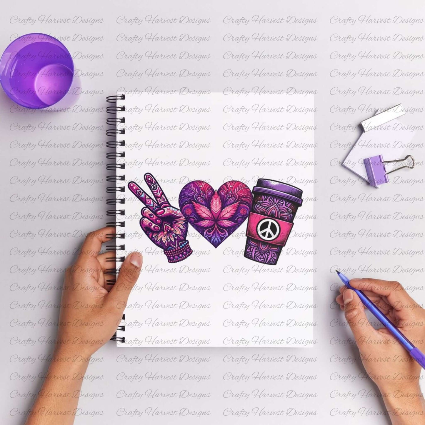 Spiral notebook featuring a peace-themed design in vibrant purple and pink tones. Includes a peace sign hand, a heart with intricate floral patterns, and a coffee cup with a peace symbol. The notebook is surrounded by purple stationery items, with hands holding a pen as if drawing or writing.