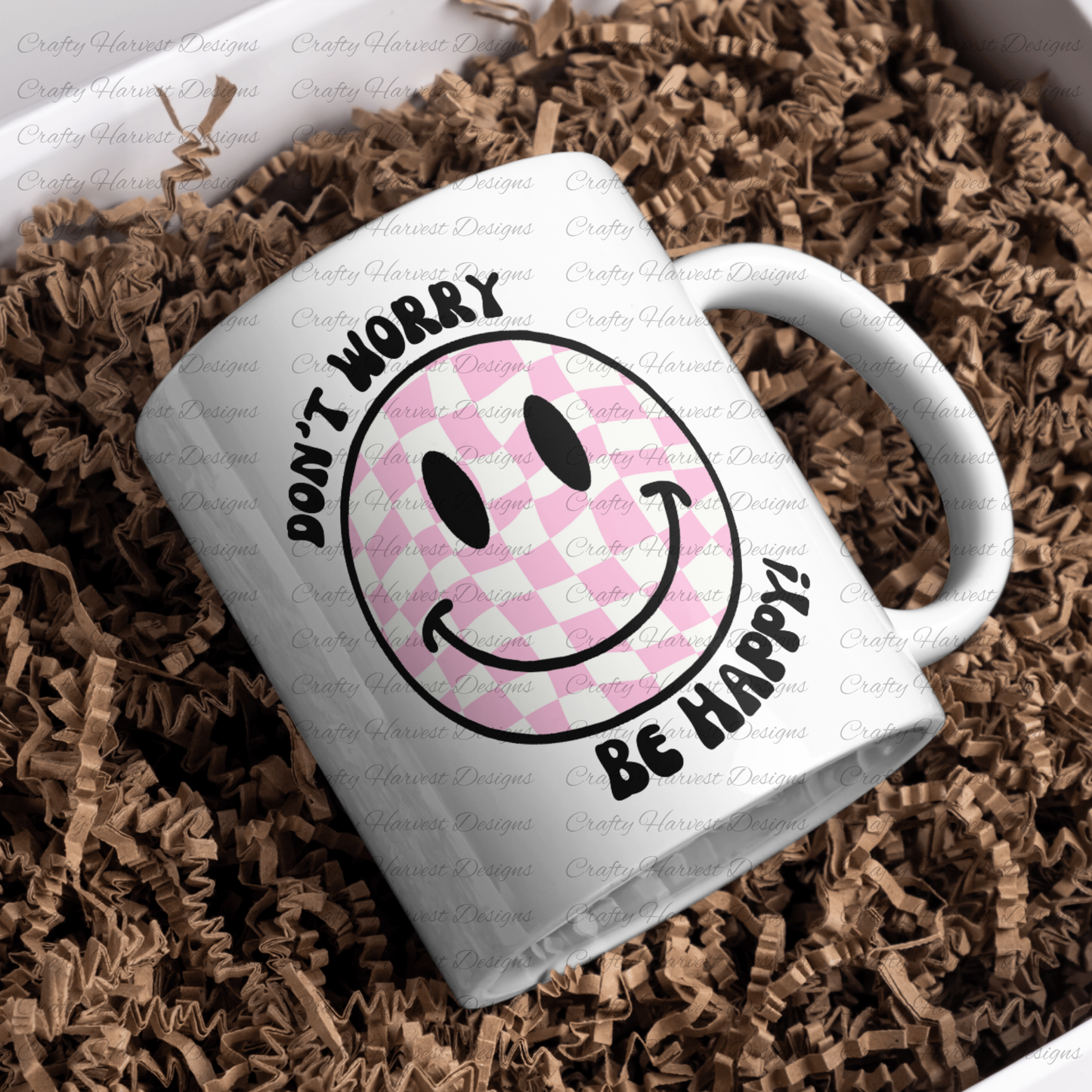 White ceramic mug featuring a pink and white checkered smiley face design with the phrase ‘Don’t Worry, Be Happy!’ in black text. The mug is placed on a bed of crinkled brown paper filler.”