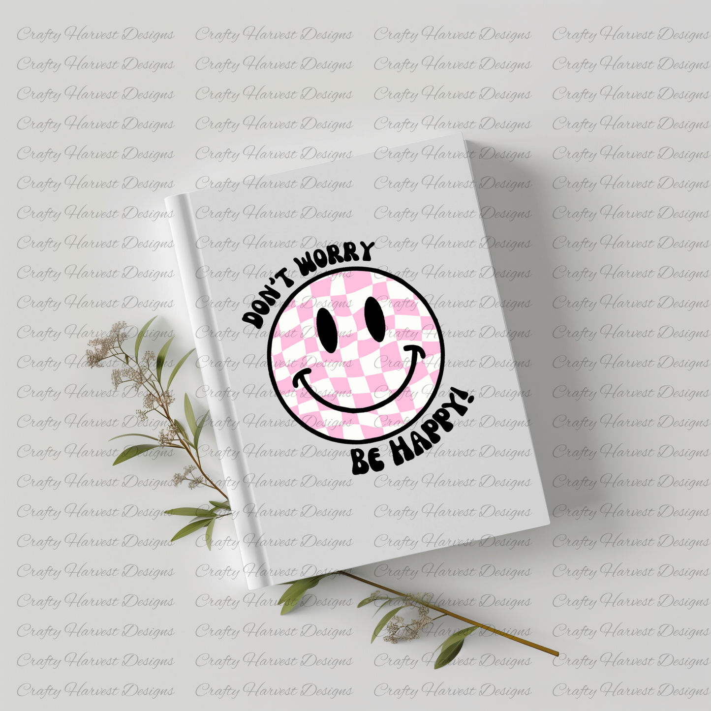 White hardcover notebook featuring a pink and white checkered smiley face design with the phrase ‘Don’t Worry, Be Happy!’ in black text. The notebook is placed on a light-colored surface with dried plant stems beside it.”
