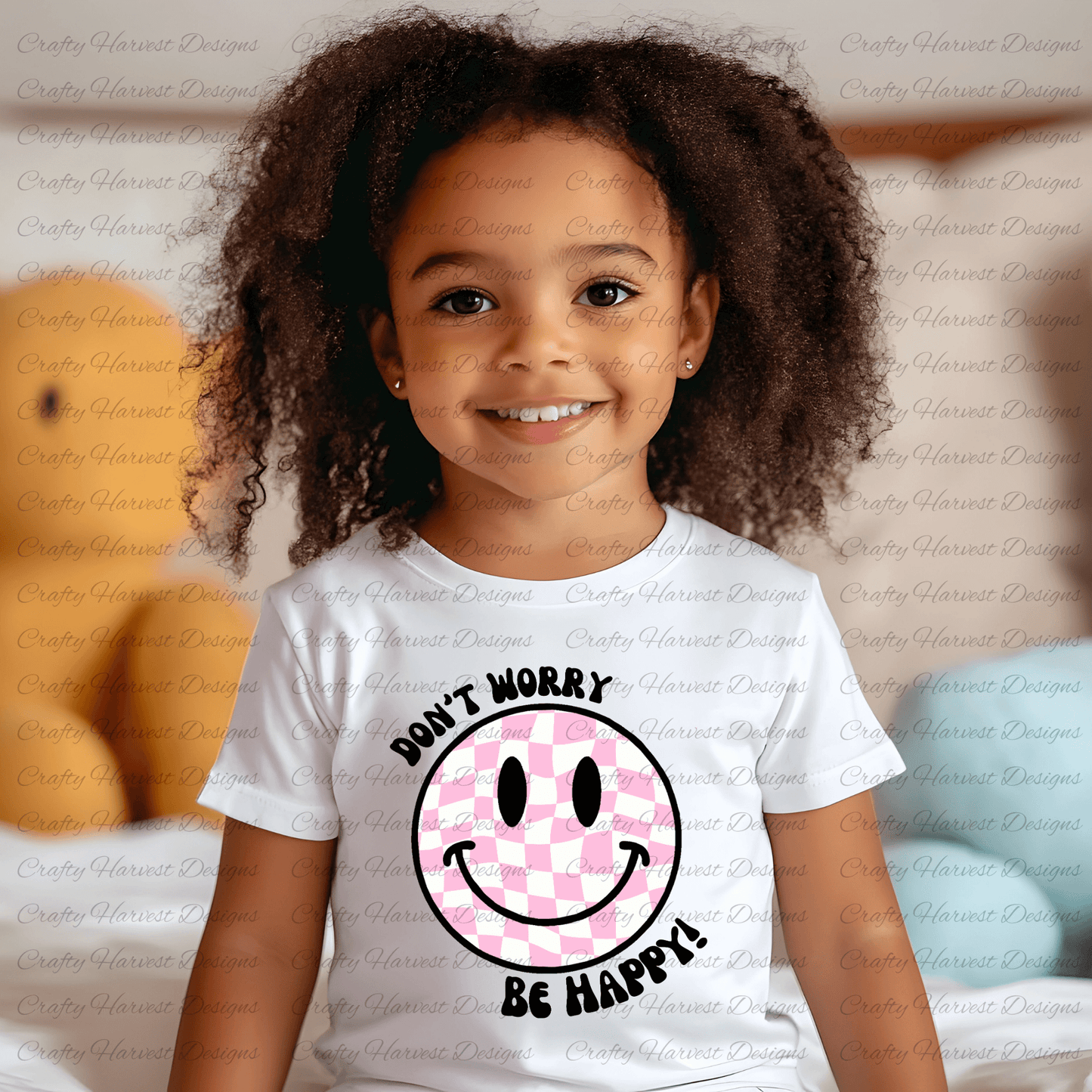 Smiling young child wearing a white t-shirt featuring a pink and white checkered smiley face design with the phrase ‘Don’t Worry, Be Happy!’ in black text. The background is softly blurred, showing a cozy indoor setting with plush toys and pillows.”