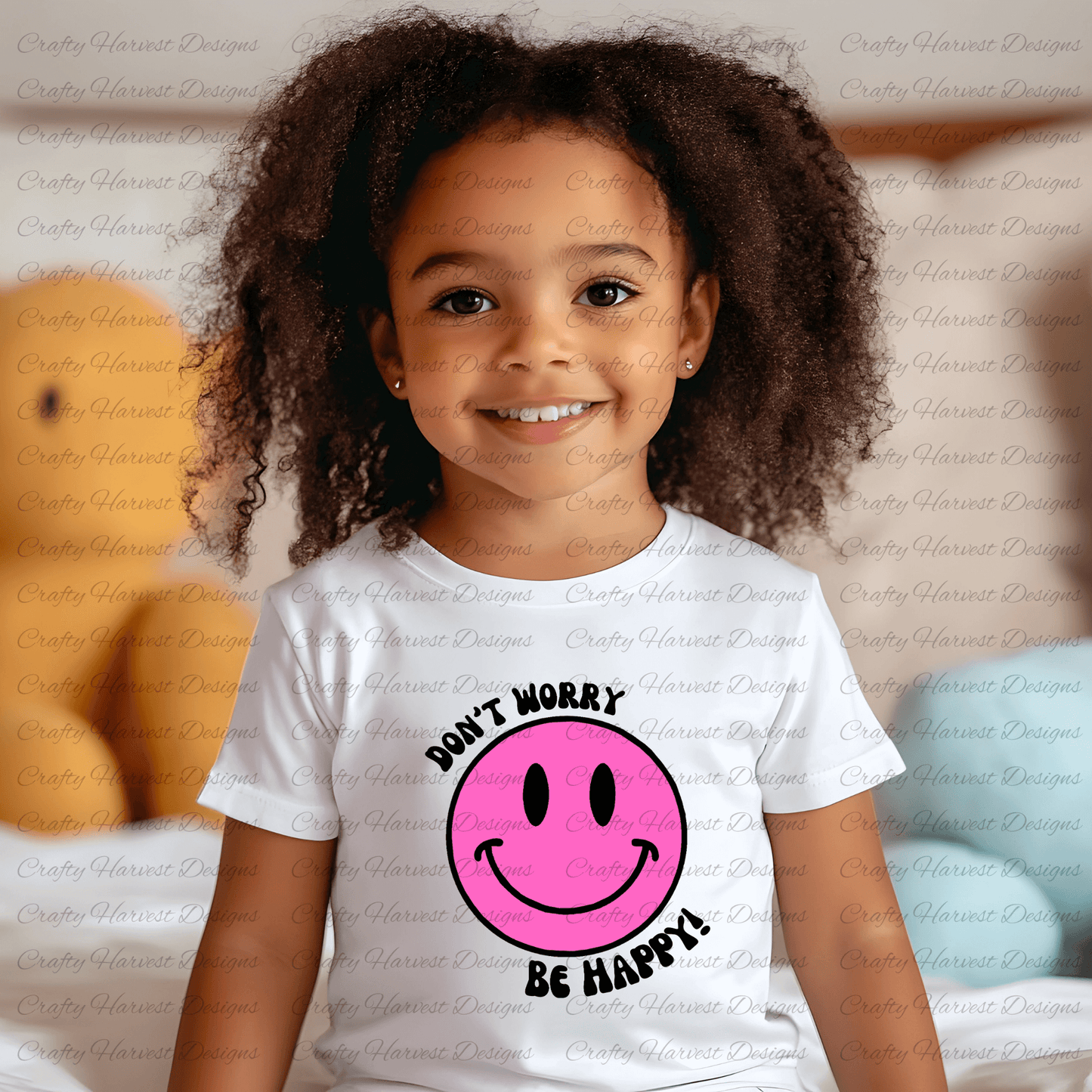 Smiling young child wearing a white t-shirt featuring a pink smiley face design with the phrase ‘Don’t Worry, Be Happy!’ in black text. The background is softly blurred, showing a cozy indoor setting with plush toys and pillows.