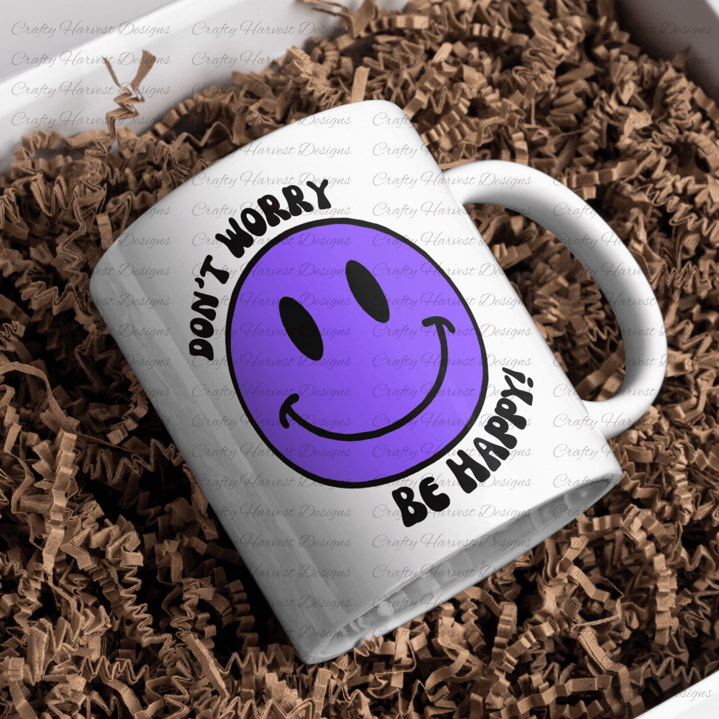White ceramic mug featuring a purple smiley face design with the phrase ‘Don’t Worry, Be Happy!’ in black text. The mug is placed on a bed of crinkled brown paper filler.