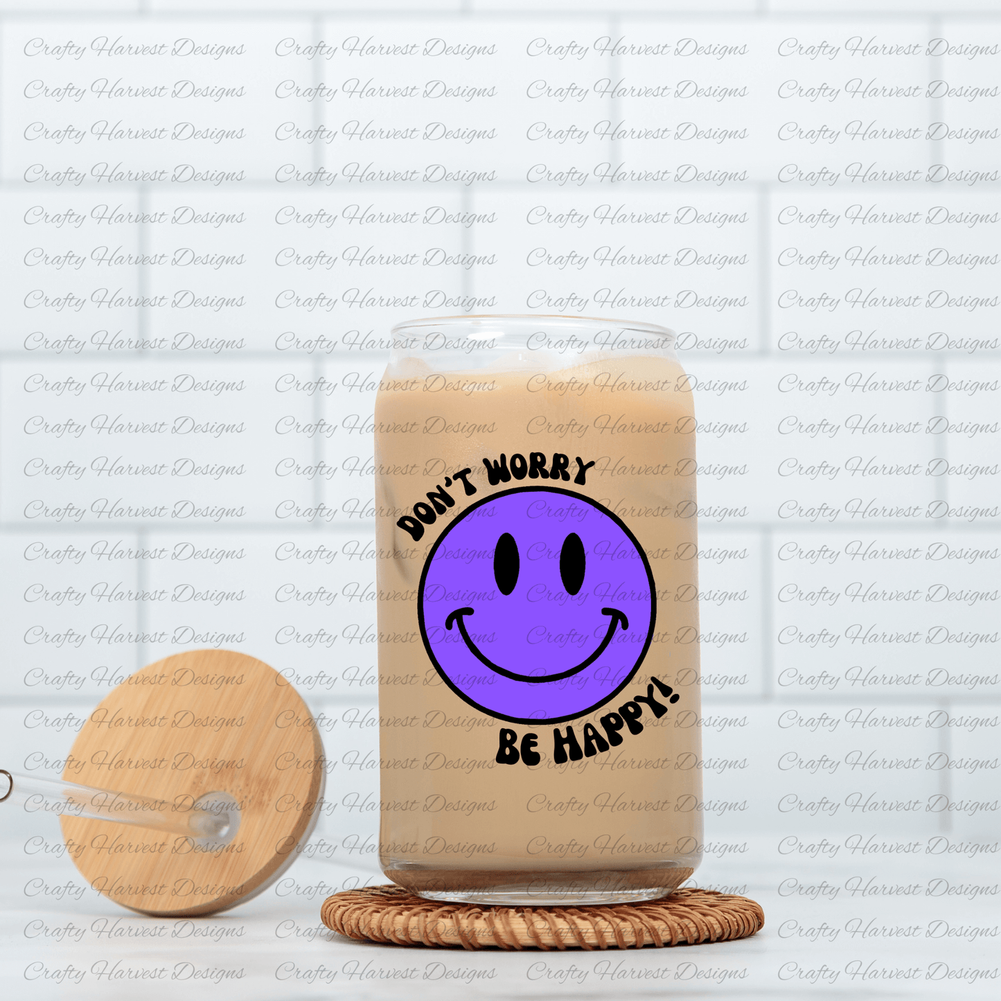Glass can tumbler filled with iced coffee, featuring a purple smiley face design with the phrase ‘Don’t Worry, Be Happy!’ in black text. The tumbler is placed on a woven coaster, with a wooden lid and glass straw resting beside it.