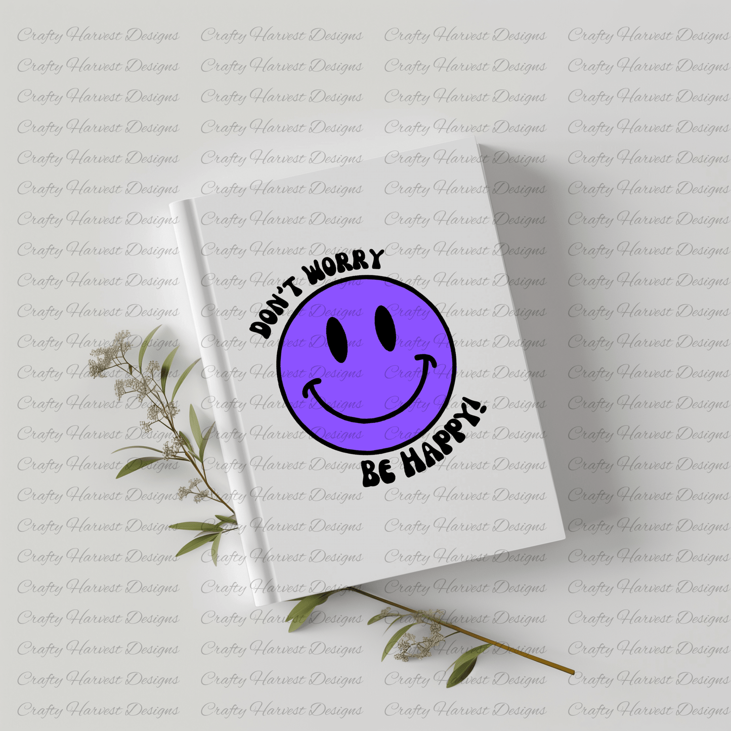 White hardcover notebook featuring a purple smiley face design with the phrase ‘Don’t Worry, Be Happy!’ in black text. The notebook is placed on a light-colored surface with dried plant stems beside it.