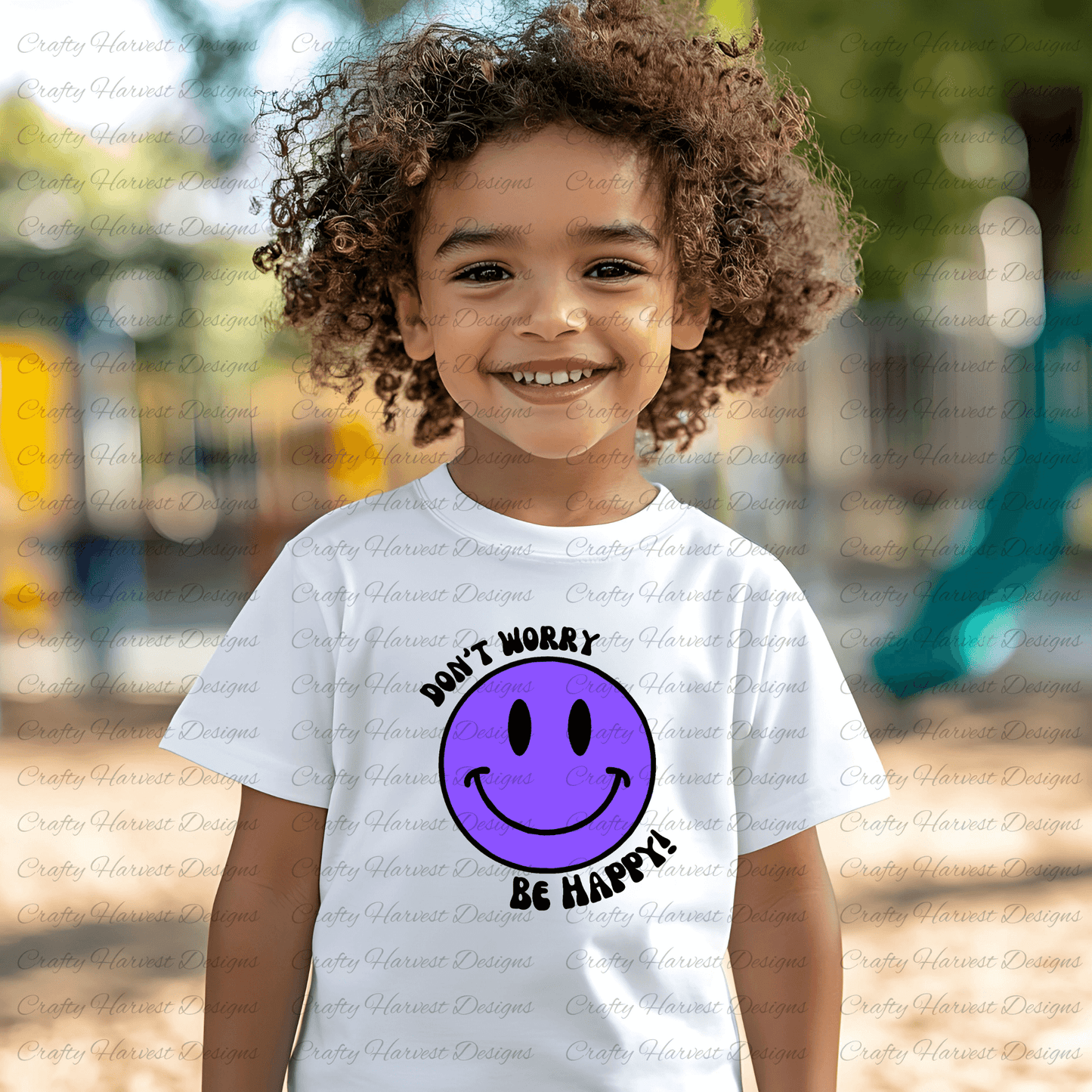 Young child wearing a white t-shirt featuring a purple smiley face with “Don’t Worry, Be Happy!” text. The child is smiling outdoors.