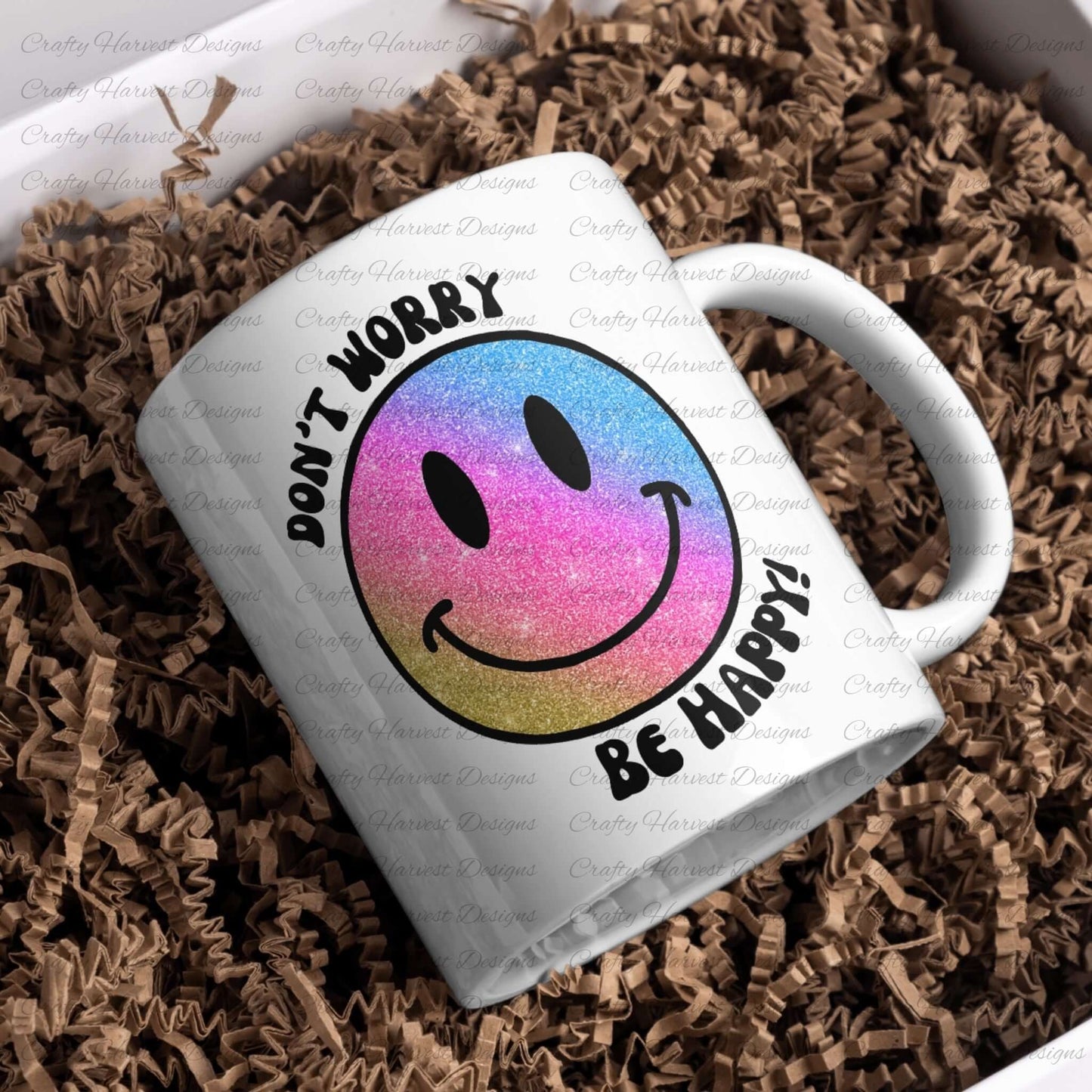 White ceramic mug featuring a glitter gradient smiley face design with the phrase ‘Don’t Worry, Be Happy!’ in black text. The smiley face transitions from pink to blue to gold. The mug is placed on a bed of crinkled brown paper filler.