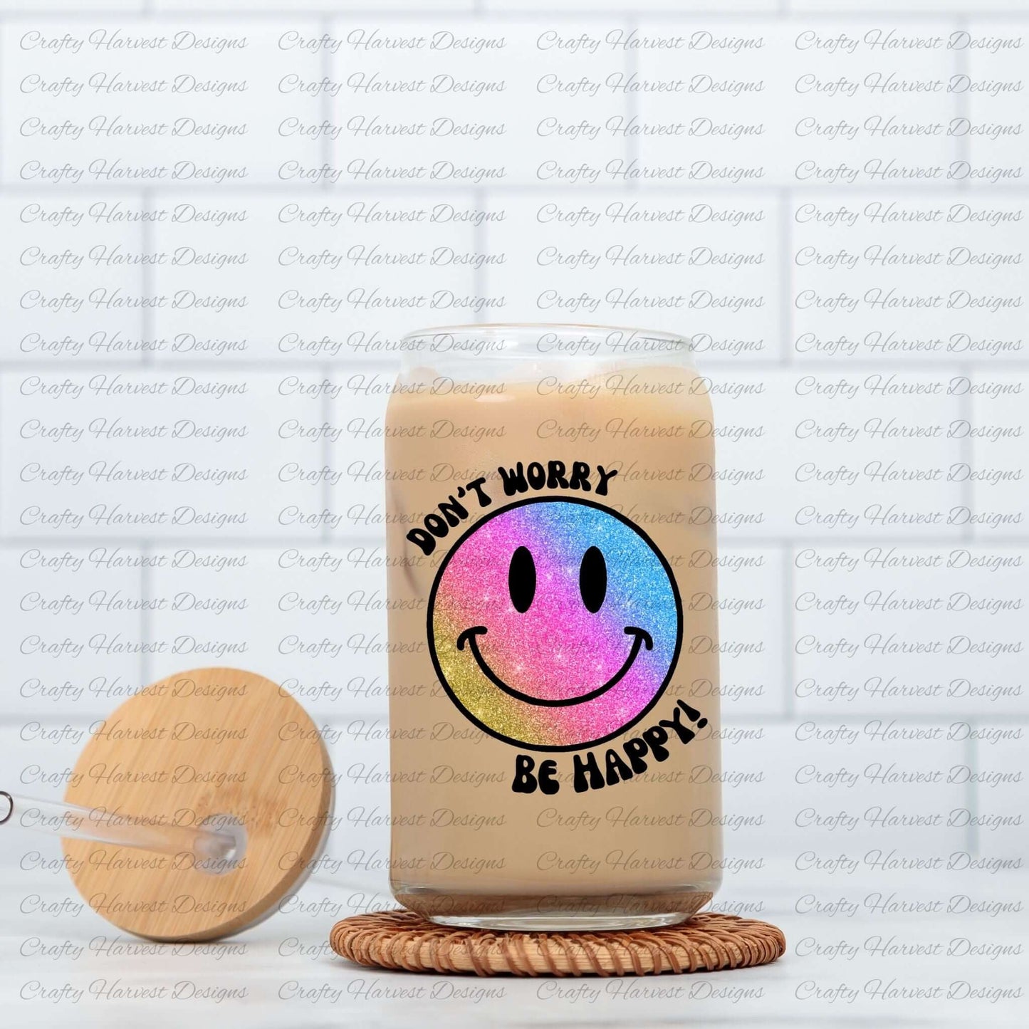 Glass can tumbler filled with iced coffee, featuring a glitter gradient smiley face design with the phrase ‘Don’t Worry, Be Happy!’ in black text. The smiley face transitions from pink to blue to gold. The tumbler is placed on a woven coaster, with a wooden lid and glass straw resting beside it.