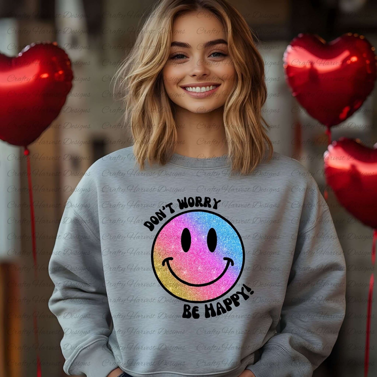 Smiling woman wearing a gray sweatshirt featuring a glitter gradient smiley face design with the phrase ‘Don’t Worry, Be Happy!’ in black text. The smiley face transitions from pink to blue to gold. She stands in front of a festive background with red heart-shaped balloons.