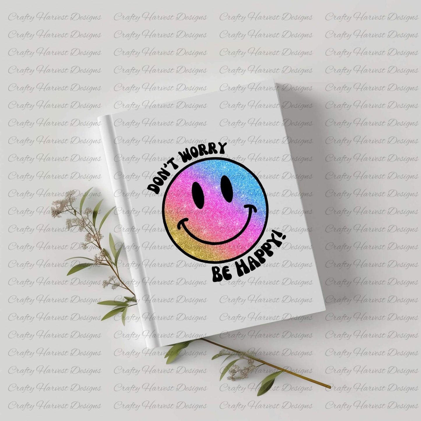 White hardcover notebook featuring a glitter gradient smiley face design with the phrase ‘Don’t Worry, Be Happy!’ in black text. The smiley face transitions from pink to blue to gold. The notebook is placed on a light-colored surface with dried plant stems beside it.