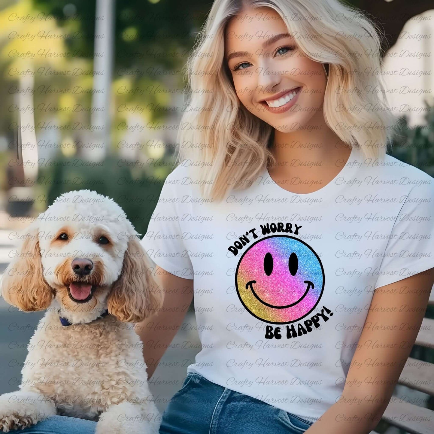 Smiling woman wearing a white t-shirt featuring a glitter gradient smiley face design with the phrase ‘Don’t Worry, Be Happy!’ in black text. The smiley face transitions from pink to blue to gold. She is outdoors with a fluffy, happy-looking dog sitting beside her.