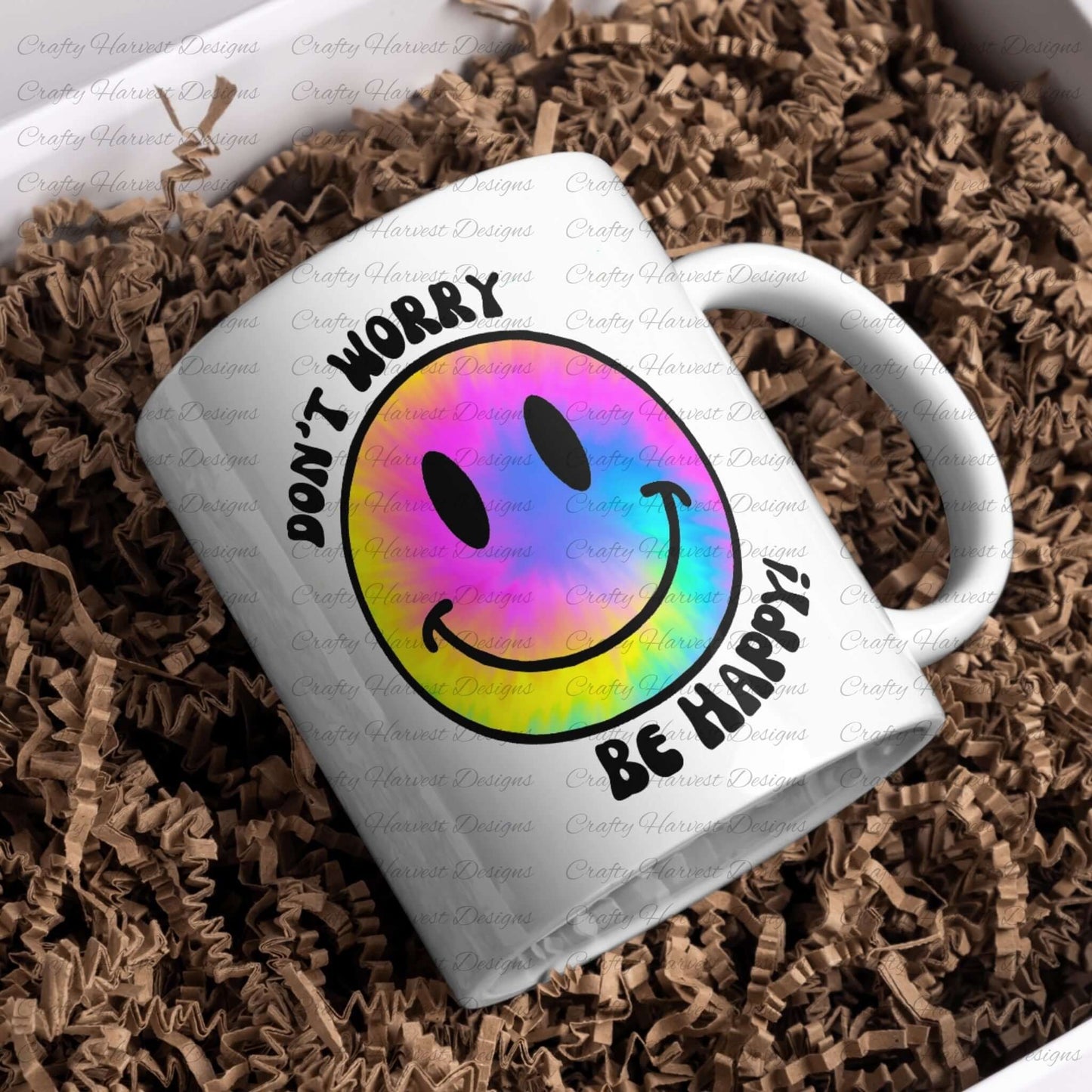 White ceramic mug featuring a rainbow tie-dye smiley face design with the phrase ‘Don’t Worry, Be Happy!’ in black text. The mug is placed on a bed of crinkled brown paper filler.