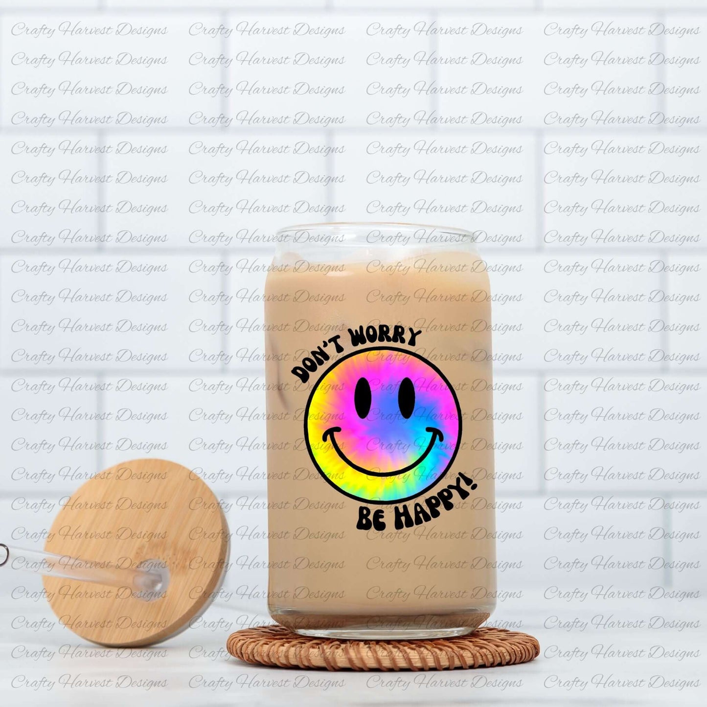 Glass can tumbler filled with iced coffee, featuring a rainbow tie-dye smiley face design with the phrase ‘Don’t Worry, Be Happy!’ in black text. The tumbler is placed on a woven coaster, with a wooden lid and glass straw resting beside it.