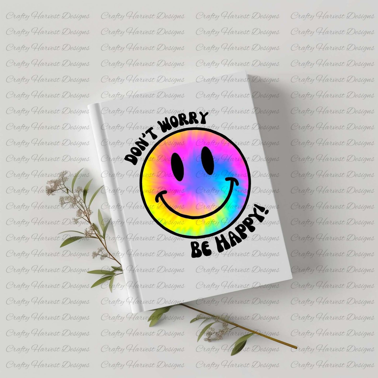 White hardcover notebook featuring a rainbow tie-dye smiley face design with the phrase ‘Don’t Worry, Be Happy!’ in black text. The notebook is placed on a light-colored surface with dried plant stems beside it.