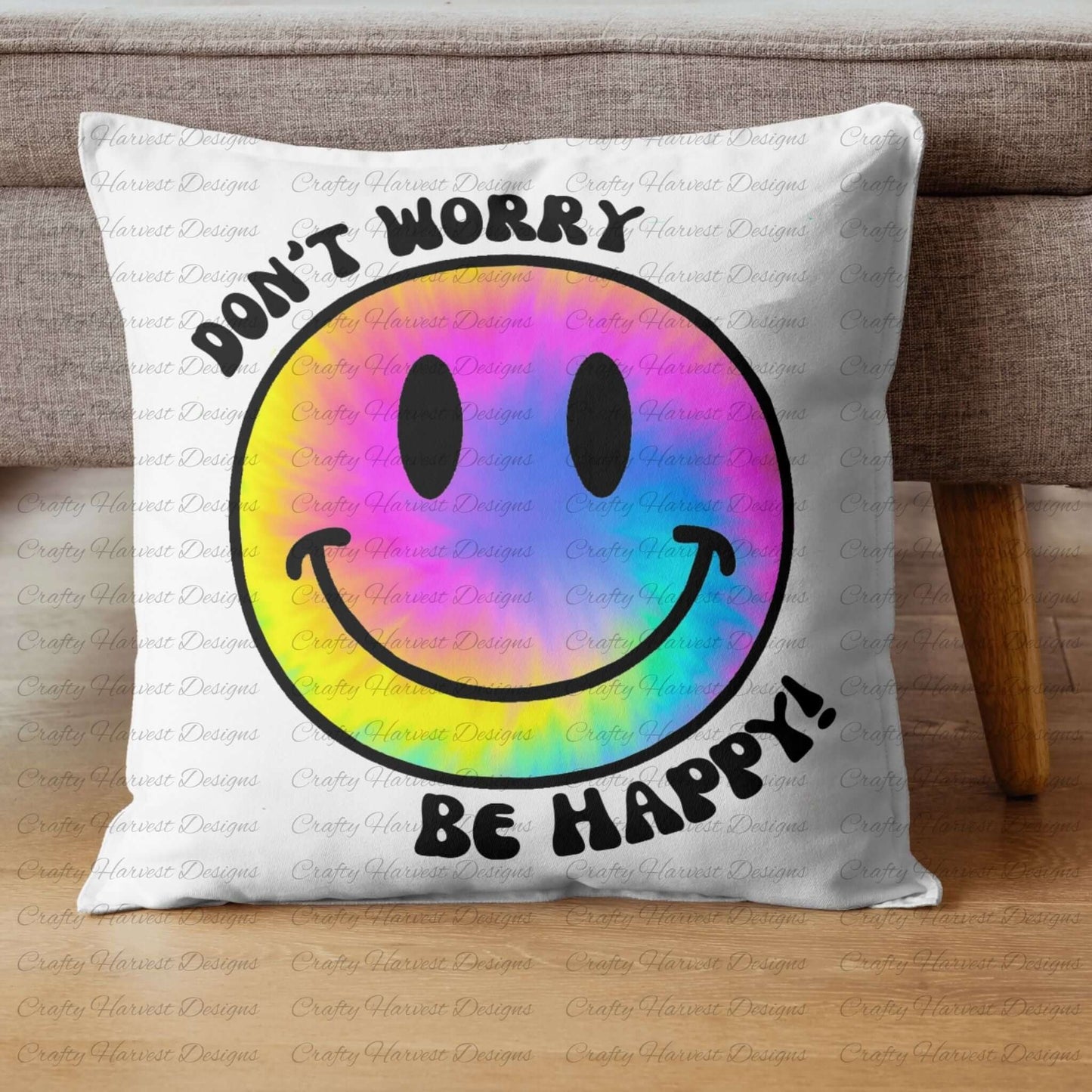 Square throw pillow resting against a couch, featuring a rainbow tie-dye smiley face design with the phrase ‘Don’t Worry, Be Happy!’ in black text.