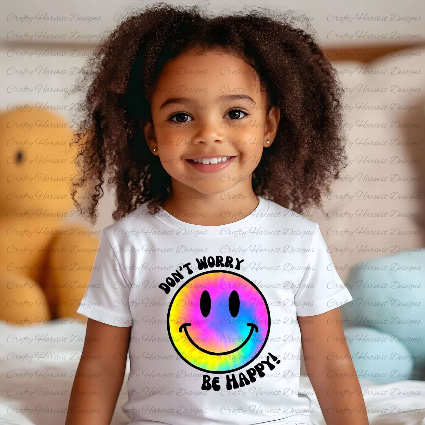 Smiling young child wearing a white t-shirt featuring a rainbow tie-dye smiley face design with the phrase ‘Don’t Worry, Be Happy!’ in black text. The background is softly blurred, showing a cozy indoor setting with plush toys and pillows.