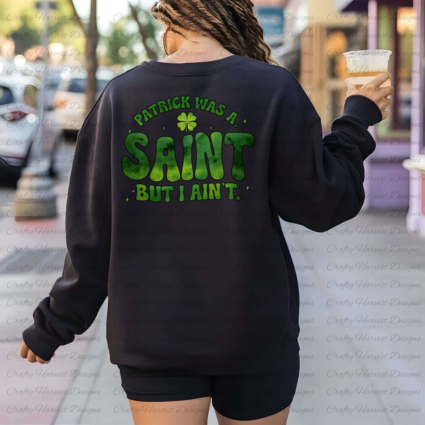 Woman walking outdoors wearing a black sweatshirt featuring the St. Patrick’s Day phrase ‘Patrick Was a Saint, But I Ain’t’ in bold green lettering with a shamrock accent. She is holding an iced coffee, showcasing the design in a casual streetwear setting. PNG and PDF download available.