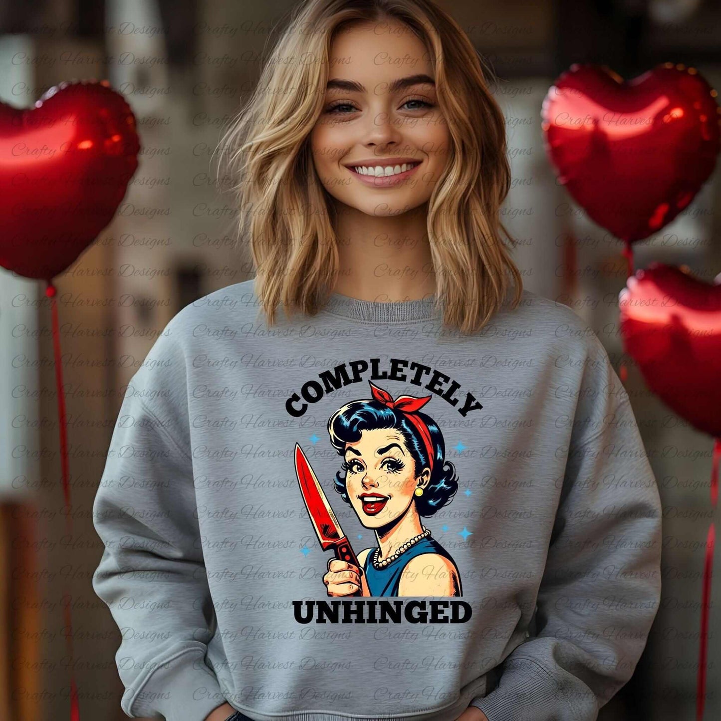 Smiling woman wearing a gray sweatshirt featuring a retro-inspired digital design of a cheerful woman holding a red-handled knife, accompanied by the bold text ‘Completely Unhinged.’ Red heart-shaped balloons are visible in the background, adding a playful and cheeky vibe to the design.