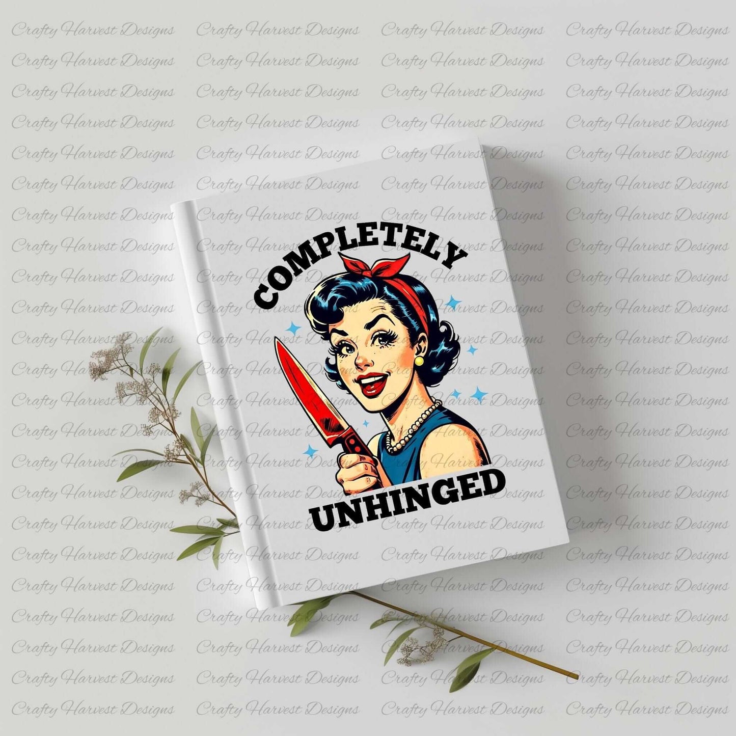 White hardcover notebook featuring a retro-inspired digital design of a cheerful woman holding a red-handled knife, accompanied by the bold text ‘Completely Unhinged.’ The notebook is displayed on a light surface with sprigs of greenery, showcasing its use as a quirky and stylish stationery item.