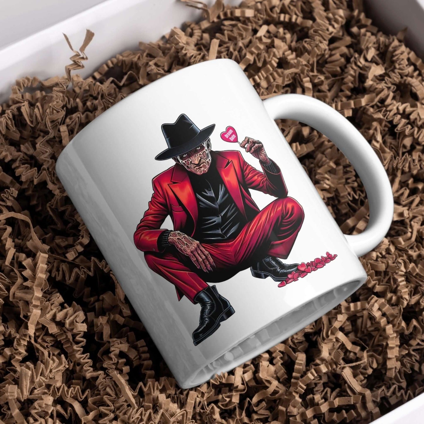 A white ceramic mug featuring a horror character design. The character is seated and dressed in a red suit with a black shirt and hat, holding a pink heart-shaped candy that reads “Be Mine.” The mug is nestled in a bed of brown crinkle paper packaging, giving a cozy and gift-ready presentation.